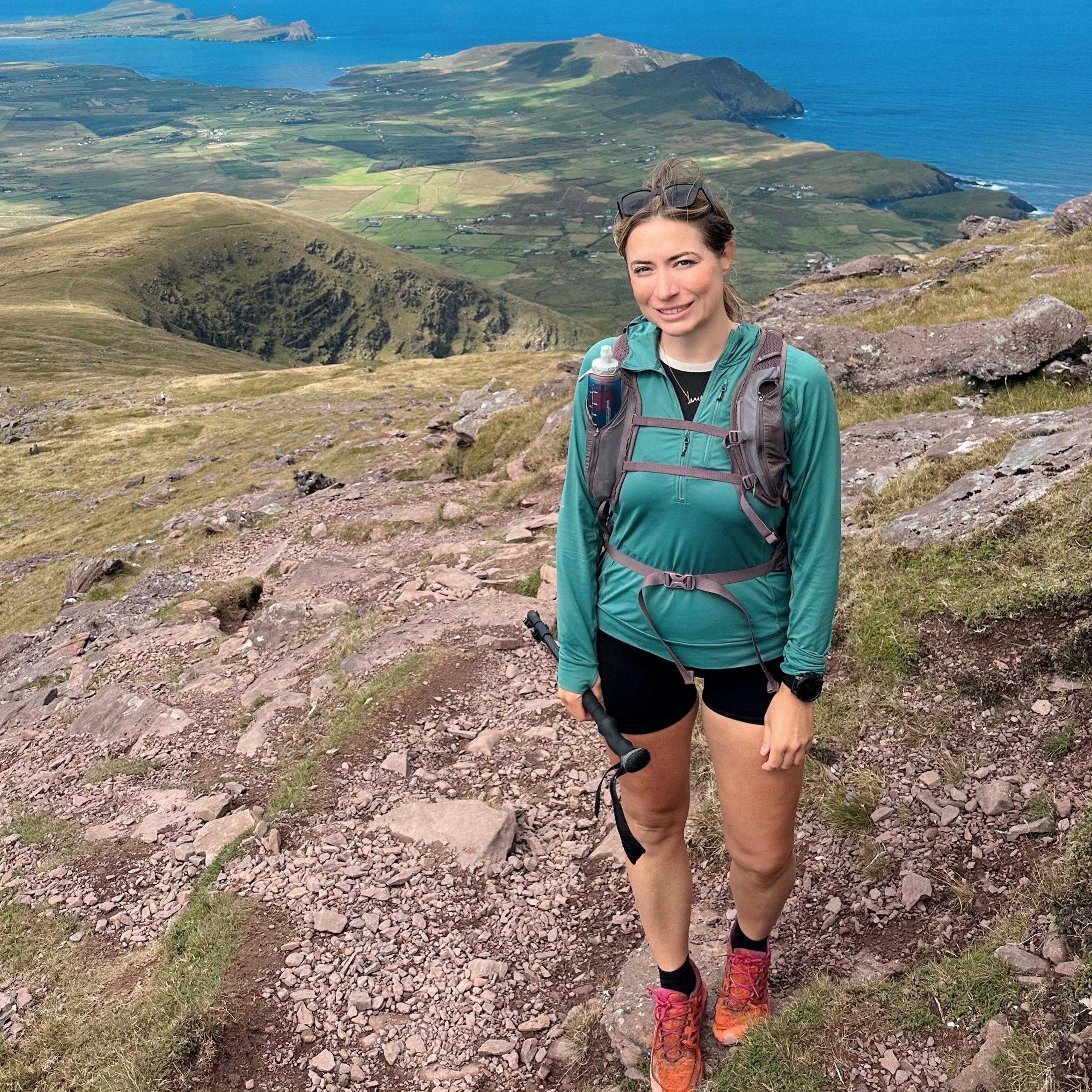 Person standing on a rocky mountain trail wearing a teal hiking top, backpack, and orange hiking shoes, holding a trekking pole. Expansive coastal landscape stretches into the distance with green fields, rugged hills, peninsulas, blue ocean, and dramatic low clouds overhead.