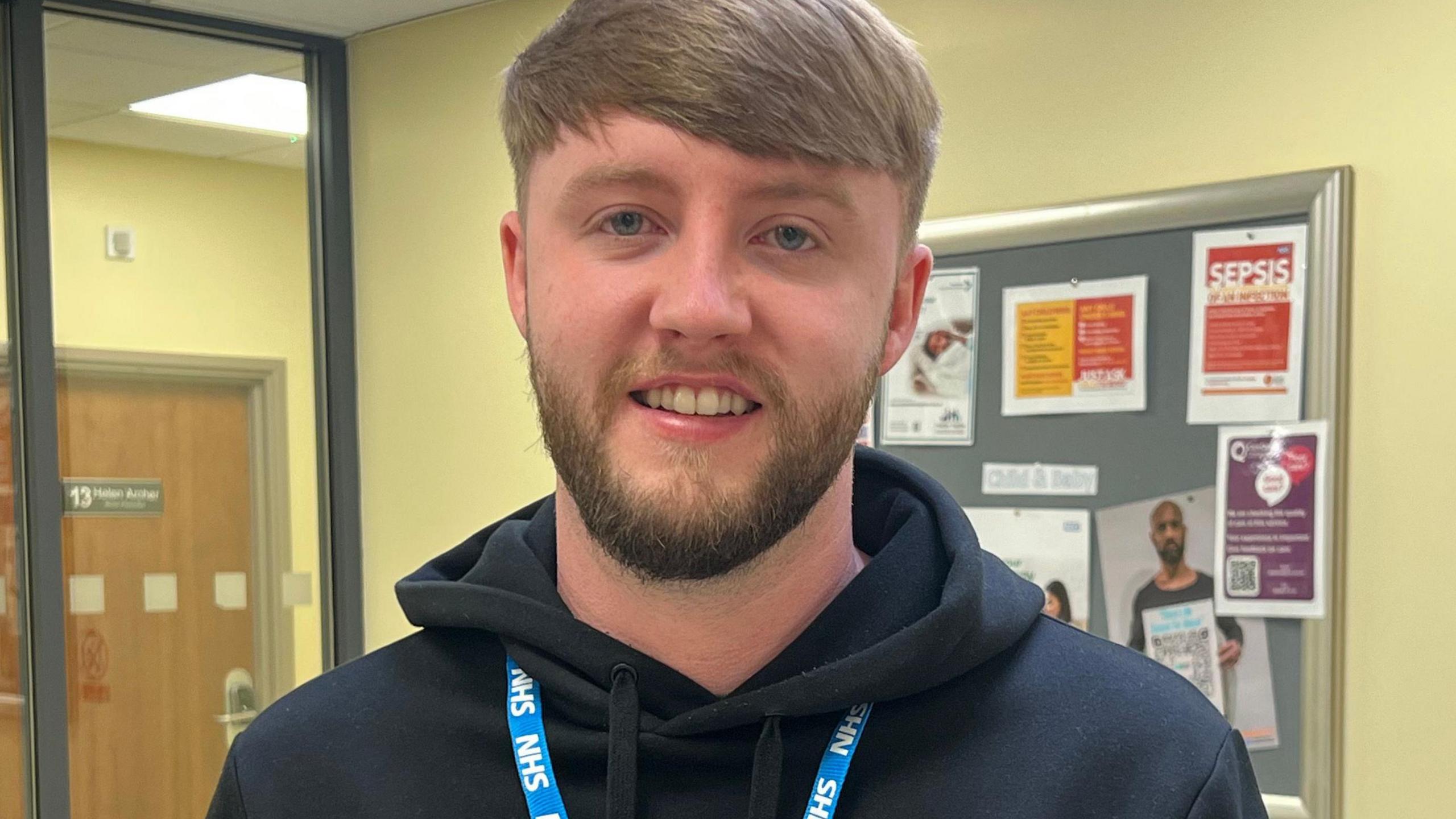 A man with short brown hair and a short beard wears a black hooded top and an NHS branded lanyard. He is standing in front of a notice board with poster on it.