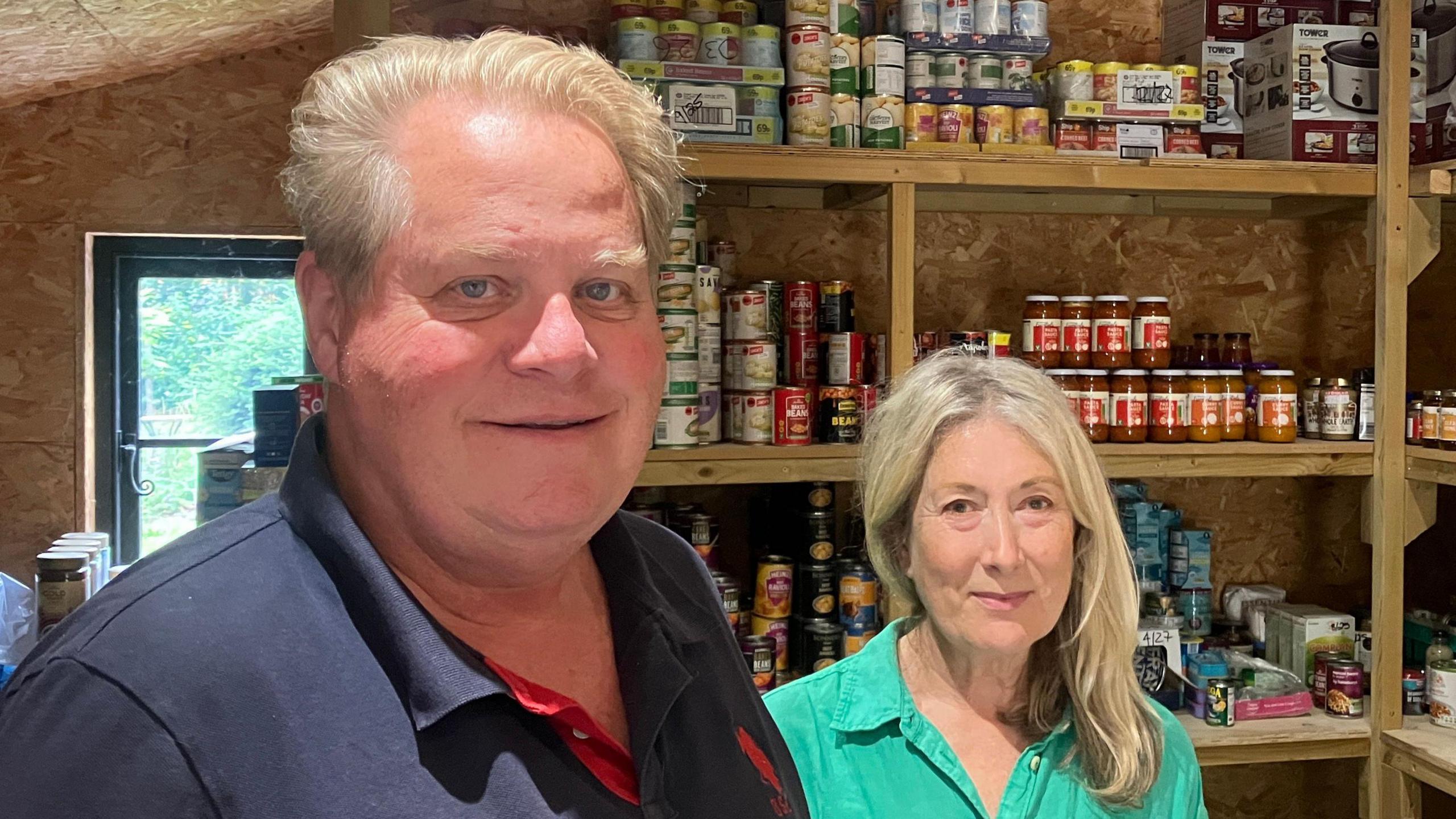 Shaun Moffat and Jo Davies smile at the camera in the Bildeston foodbank. Shaun has short grey hair and wears a blue polo top. Jo has long blonde hair and wears a green shirt.