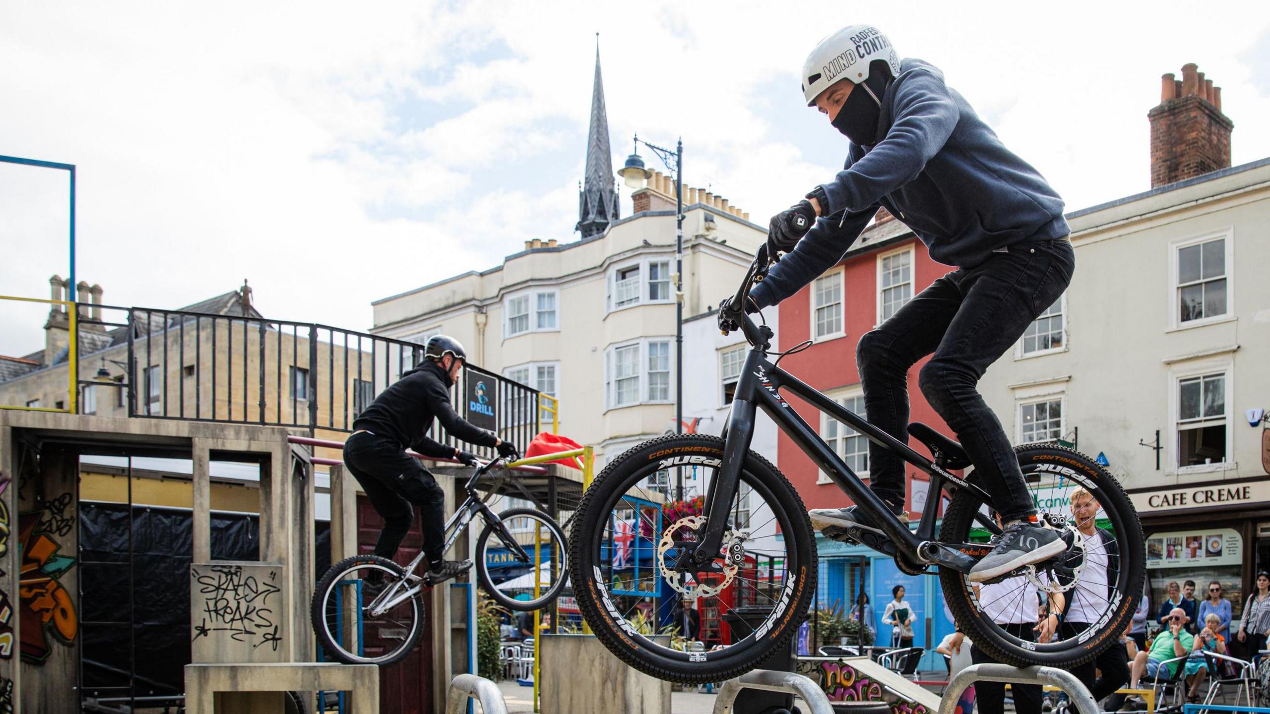 Bike riders performing stunts - two cyclists are riding over obstacles with shops and onlookers in the background