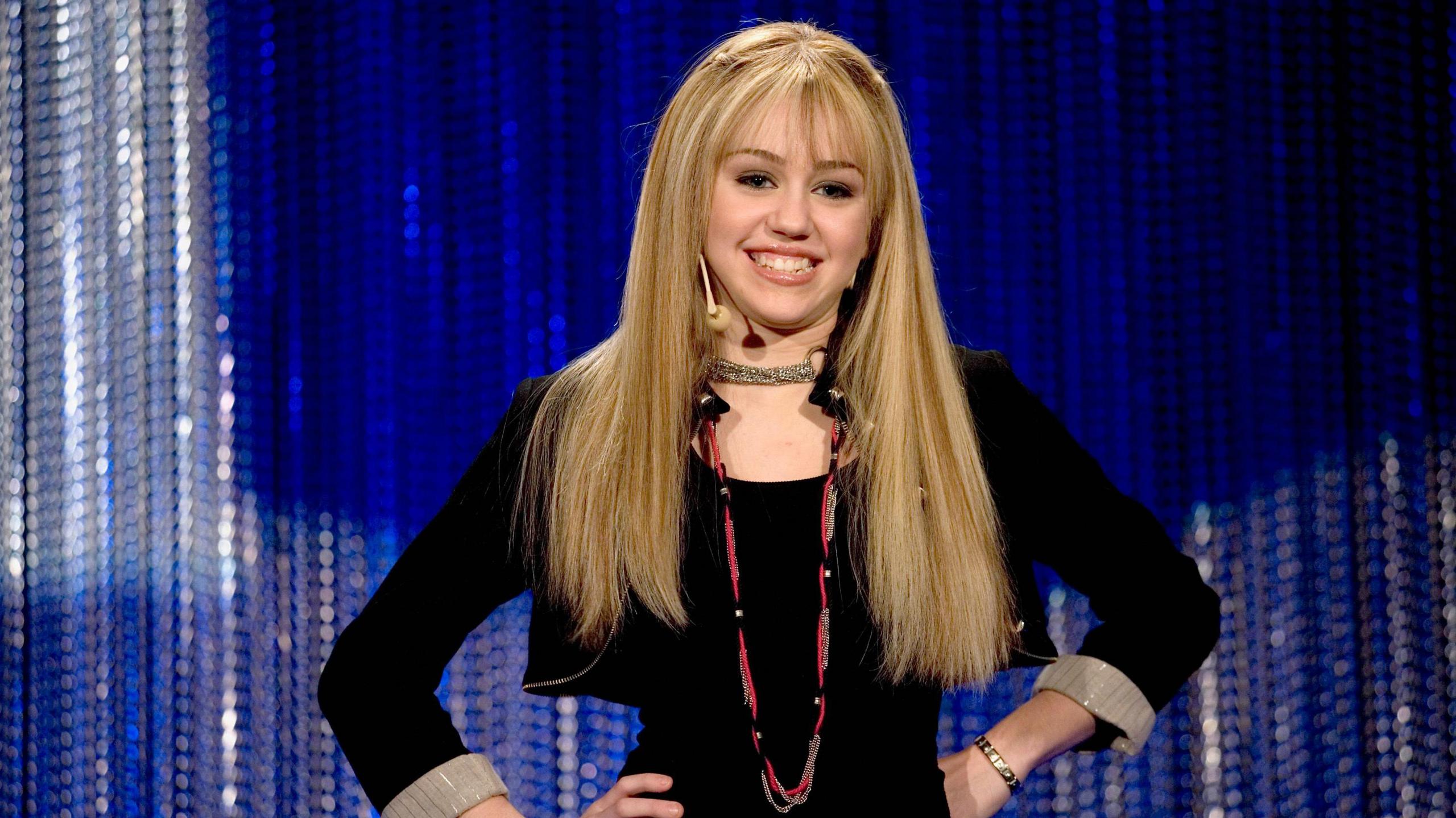 Miley Cyrus as Hannah Montana. Hannah is a teenage pop star, she wears a long blonde wig and black jacket over a black top, posing with her hands on her hips.  