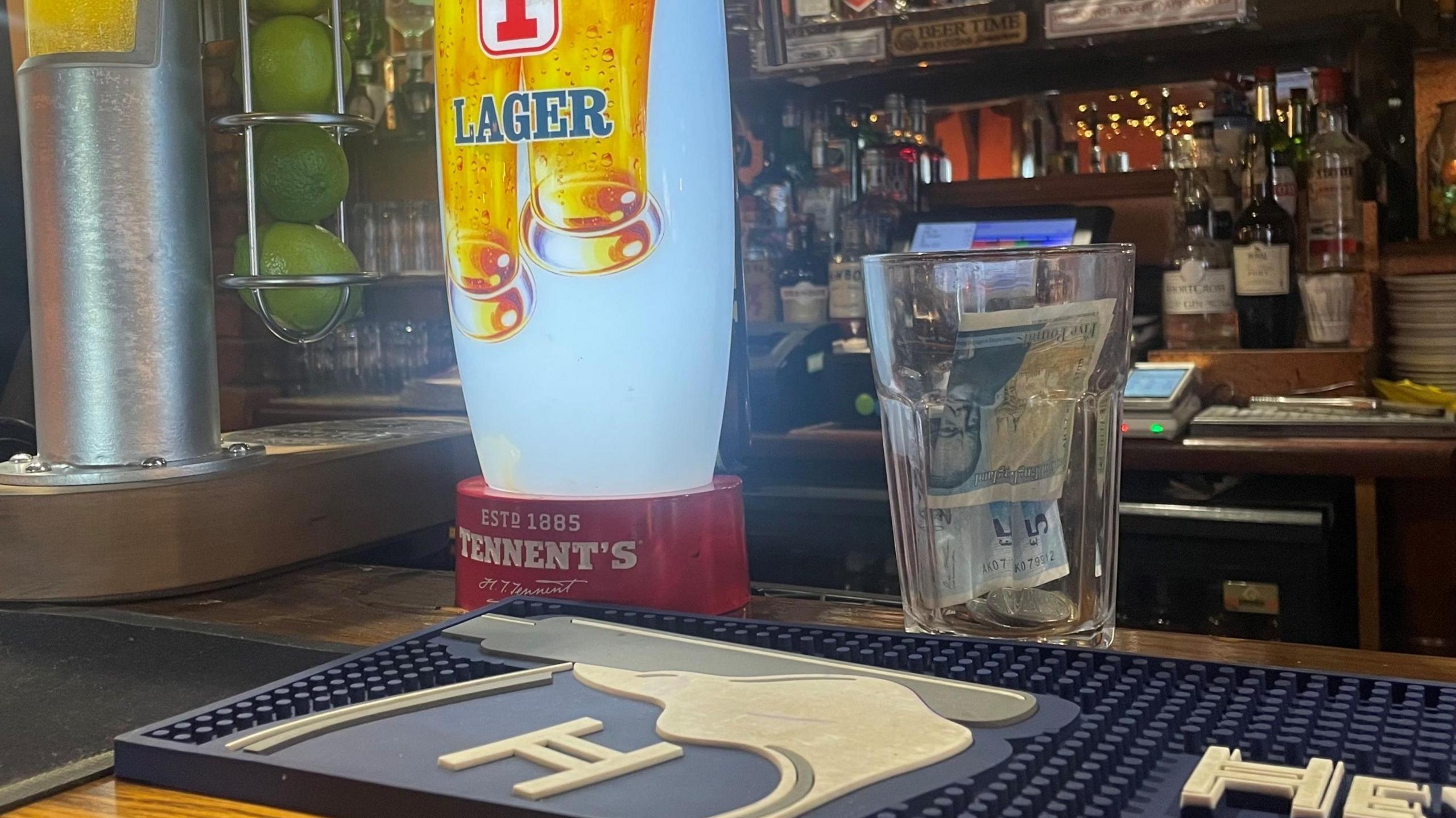 A clear glass with a 5 pound note and several coins sits beside a beer tap on top of a bar