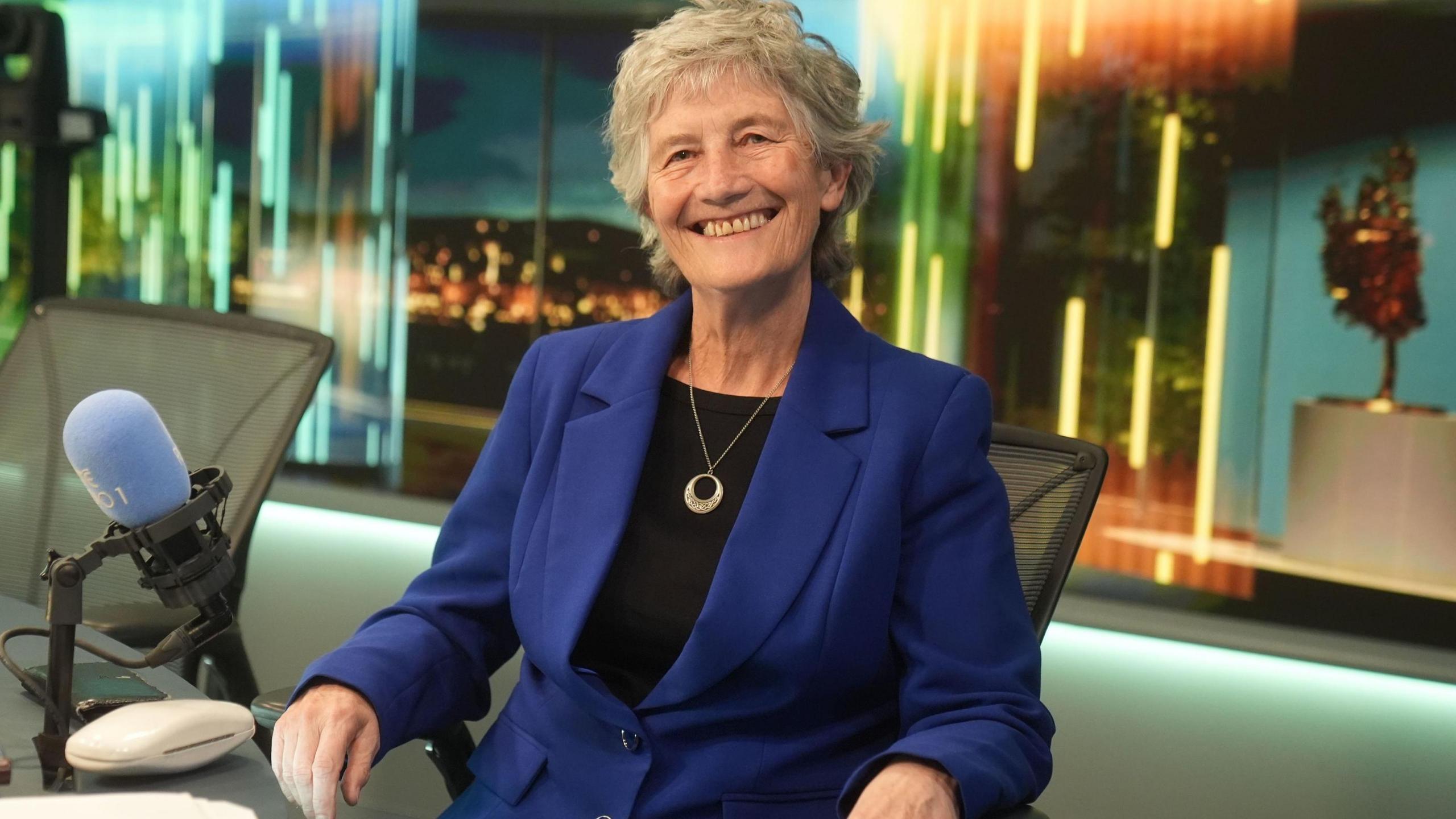 Catherine Connolly has grey hair and is smiling broadly. She is wearing a blue jacket and black blouse. She is sitting beside a radio studio microphone.