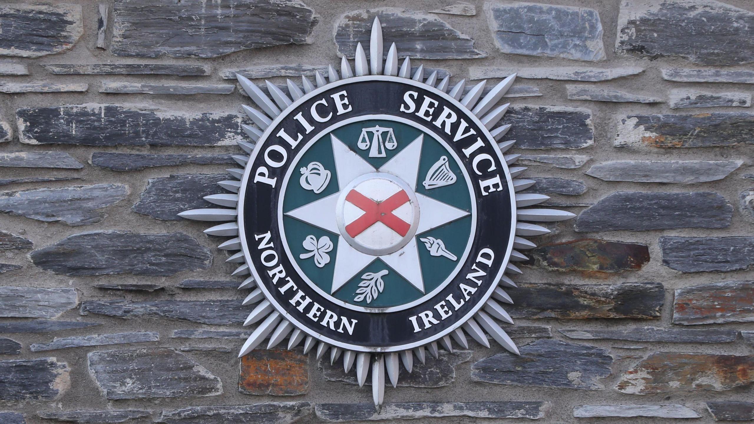 PSNI logo is on a mixed stone wall. The logo has a red and white cross centre, with a six pronged star surrounding it, there are six silver logos on a green background. There is an outer circle of silver writing on a black background. It says Police Service Northern Ireland. Around the entire logo are silver spikes.  