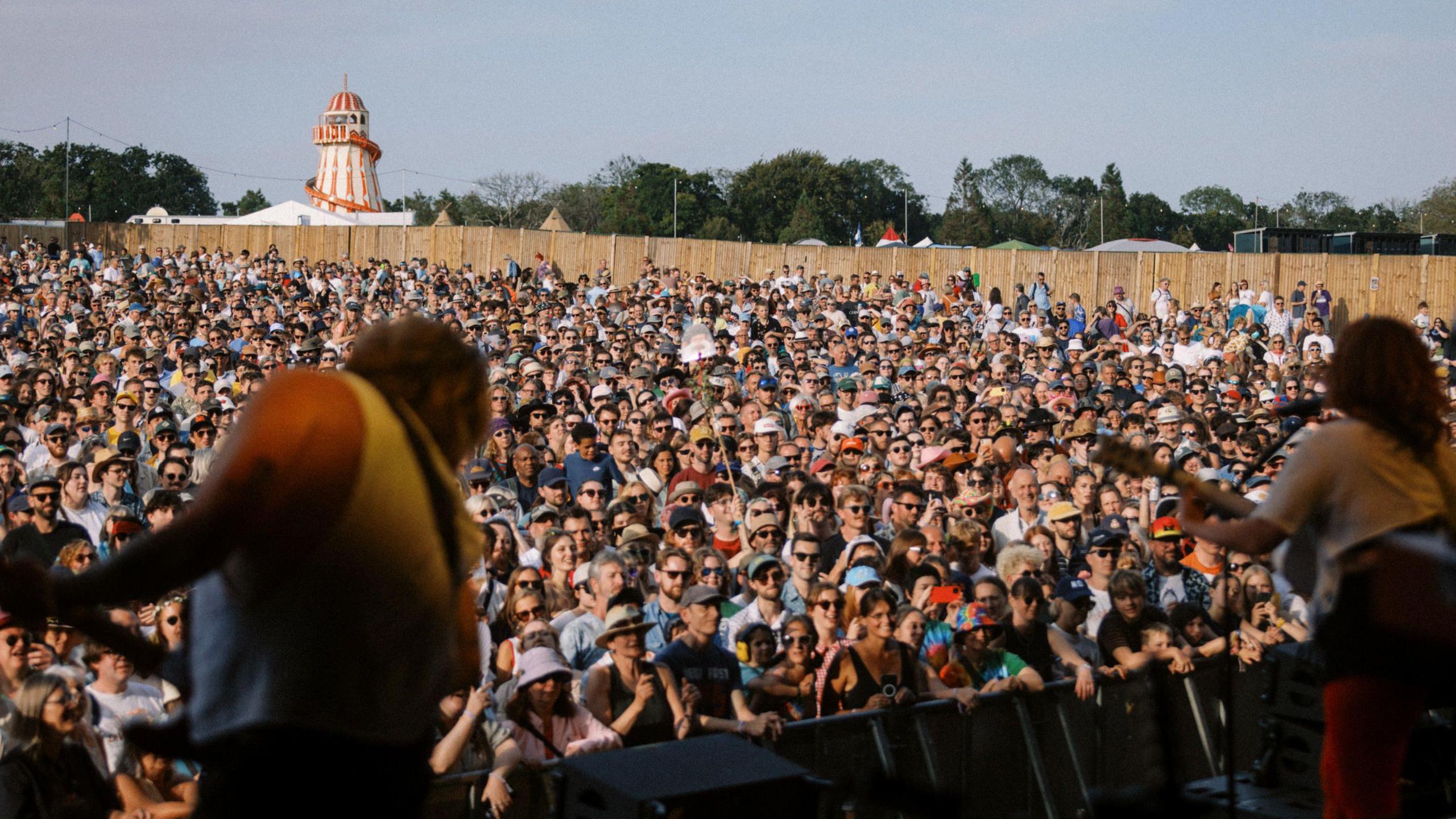 A photo of the large festival crowd taken from the stage, with two men playing guitars blurred in the foreground. Behind the crowd is a line of trees and a helter skelter.