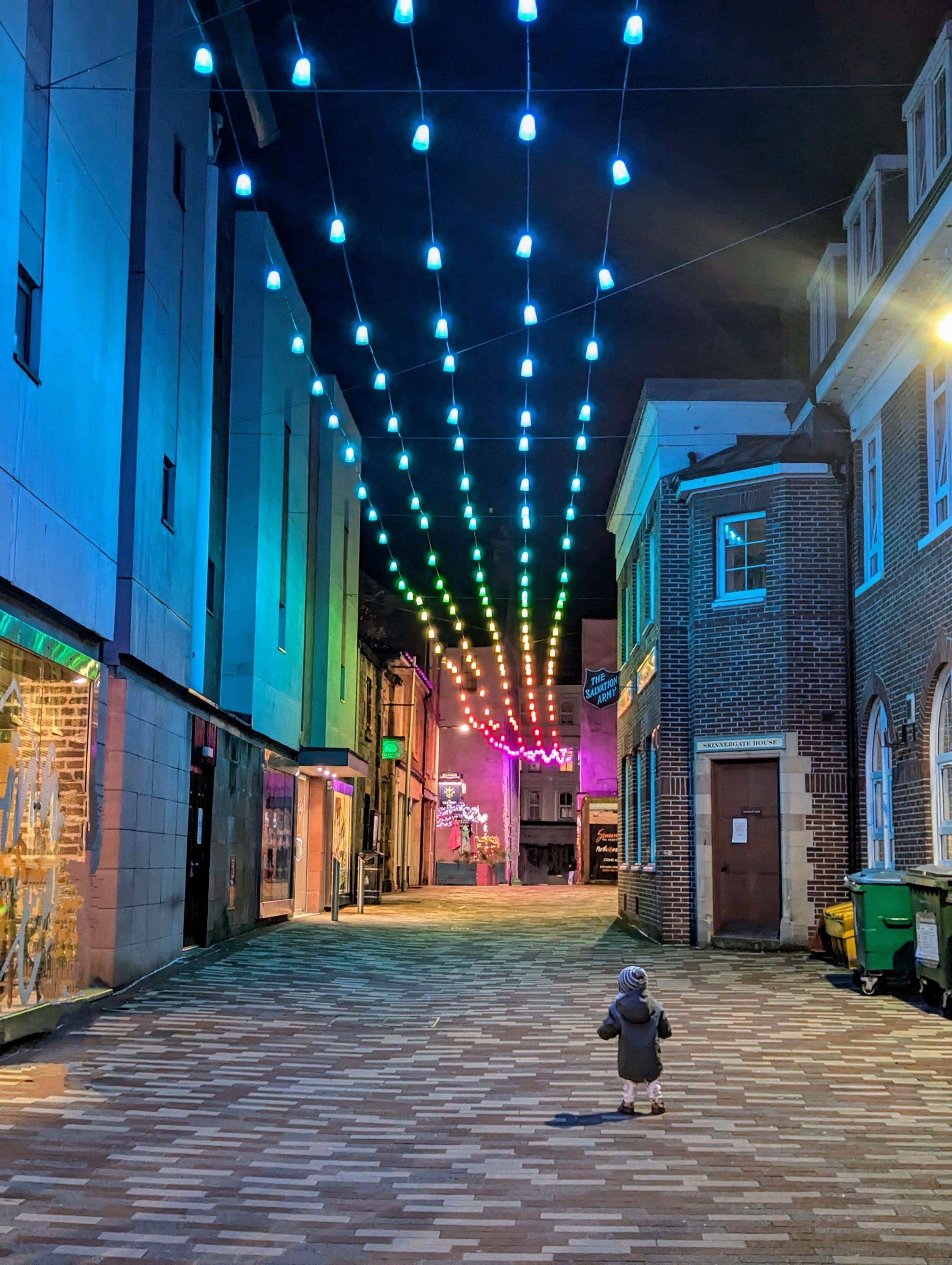 A small boy on a cobbled street stares up at colourful lights strung up between an alleyway of buildings