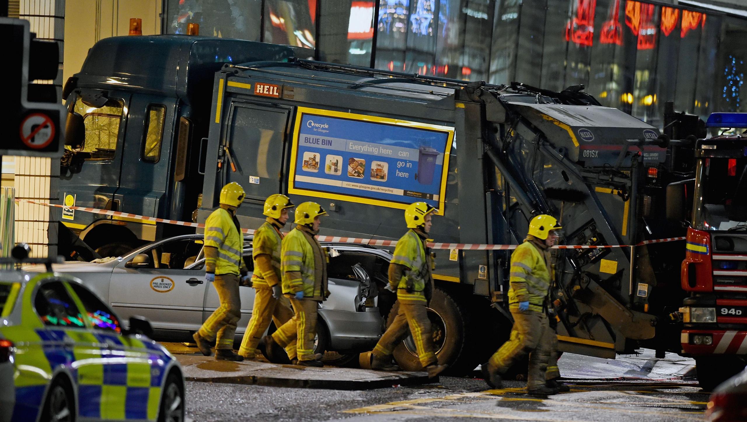 Five firefighters walk past the Glasgow City Council bin lorry, which has come to rest at the side of the Millennium Hotel in George Square. The vehicle, which crashed into a grey taxi, is cordoned off with red and white tape. A parked police car is also visible in the image.