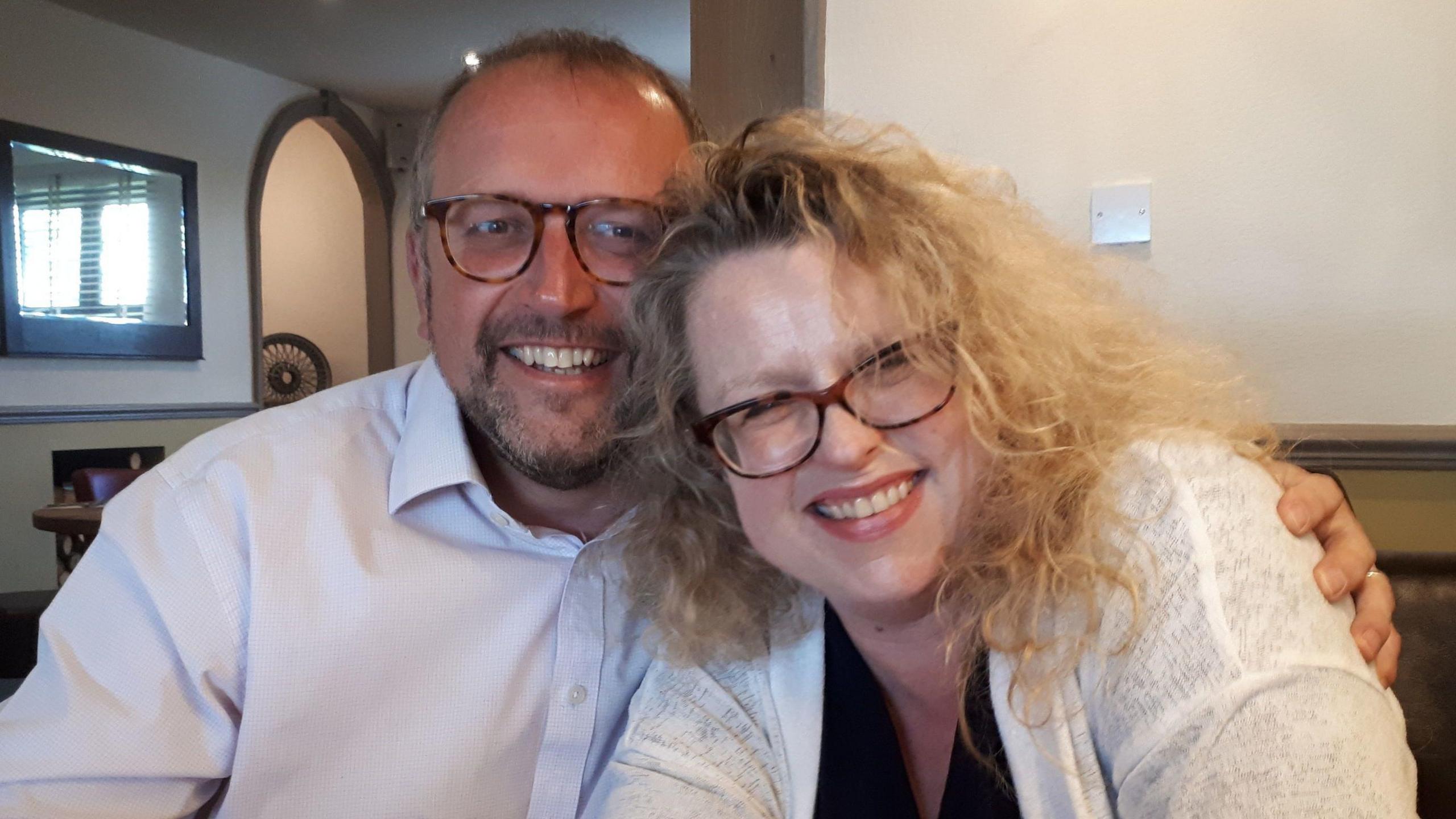 Two people are smiling at the camera. On the left is Rich Williman, who has a white shirt on and is wearing glasses. He has his arm around Erika Williman, who has blonde curly hair and is also wearing glasses.