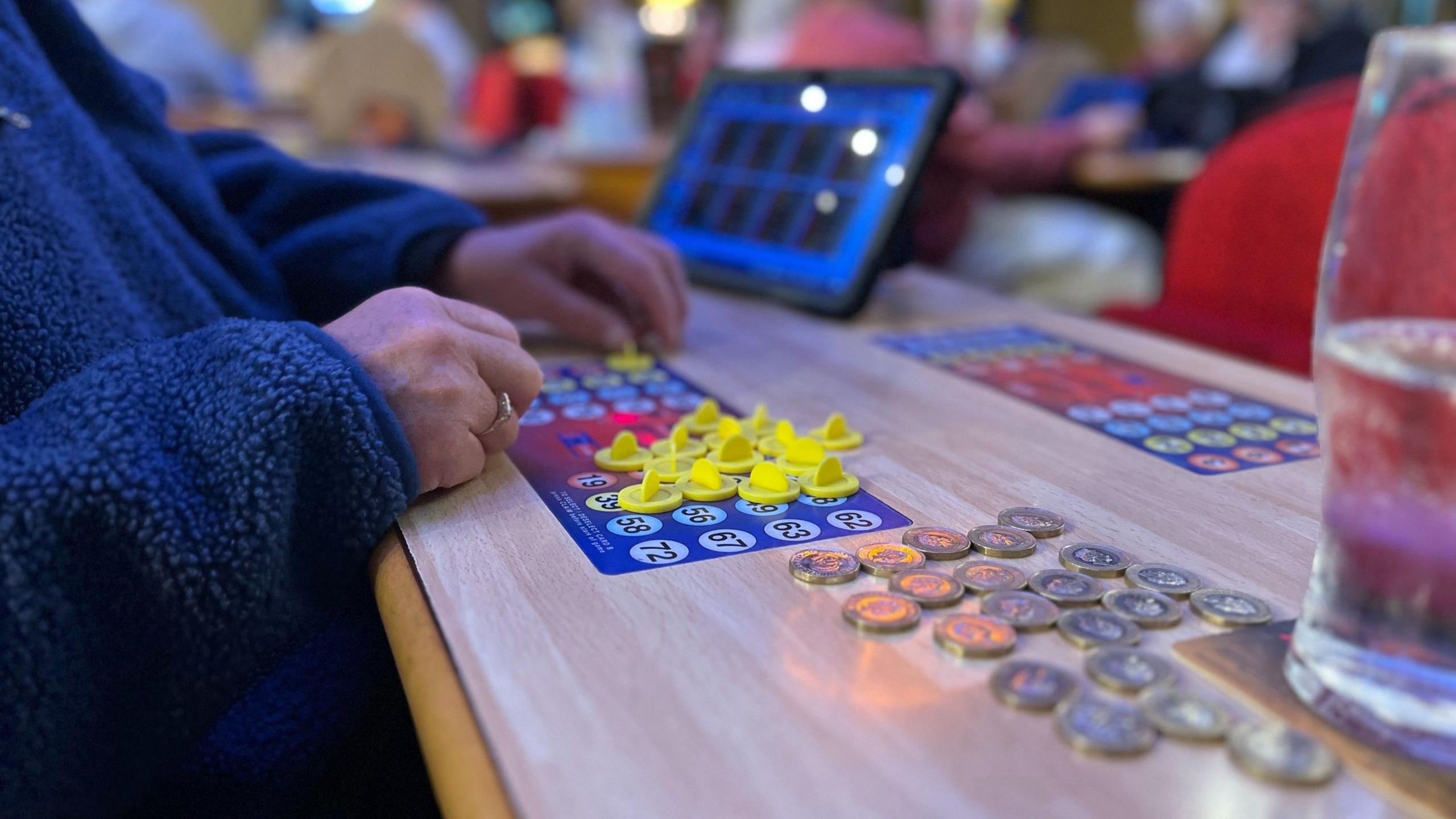 A woman wearing a blue fleece moves bingo counters about on a board, with some pound coins on the table next to her.