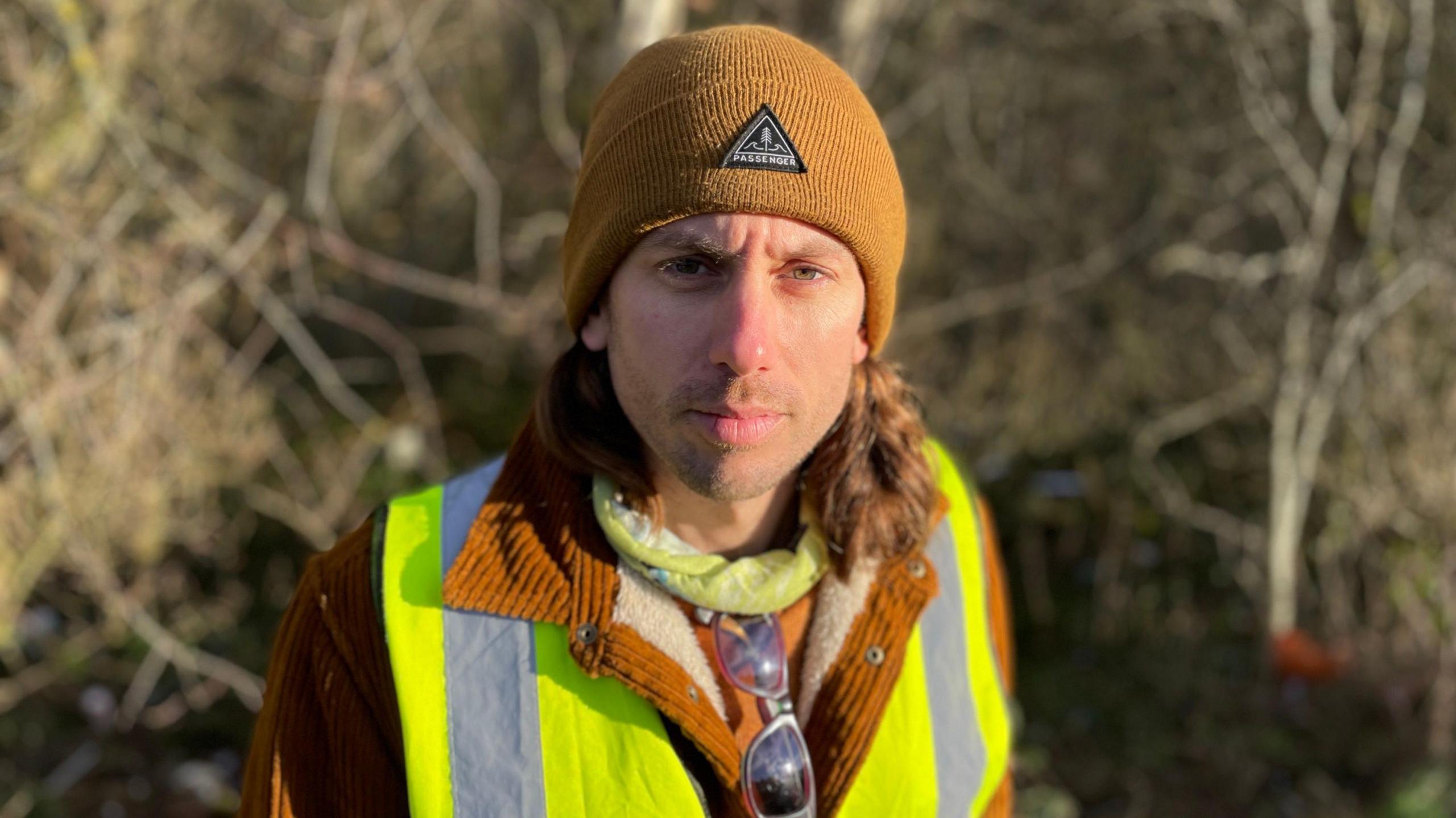 A man with a brown beanie hat, brown coat and hi-vis jacket looks at the camera, with trees in the background