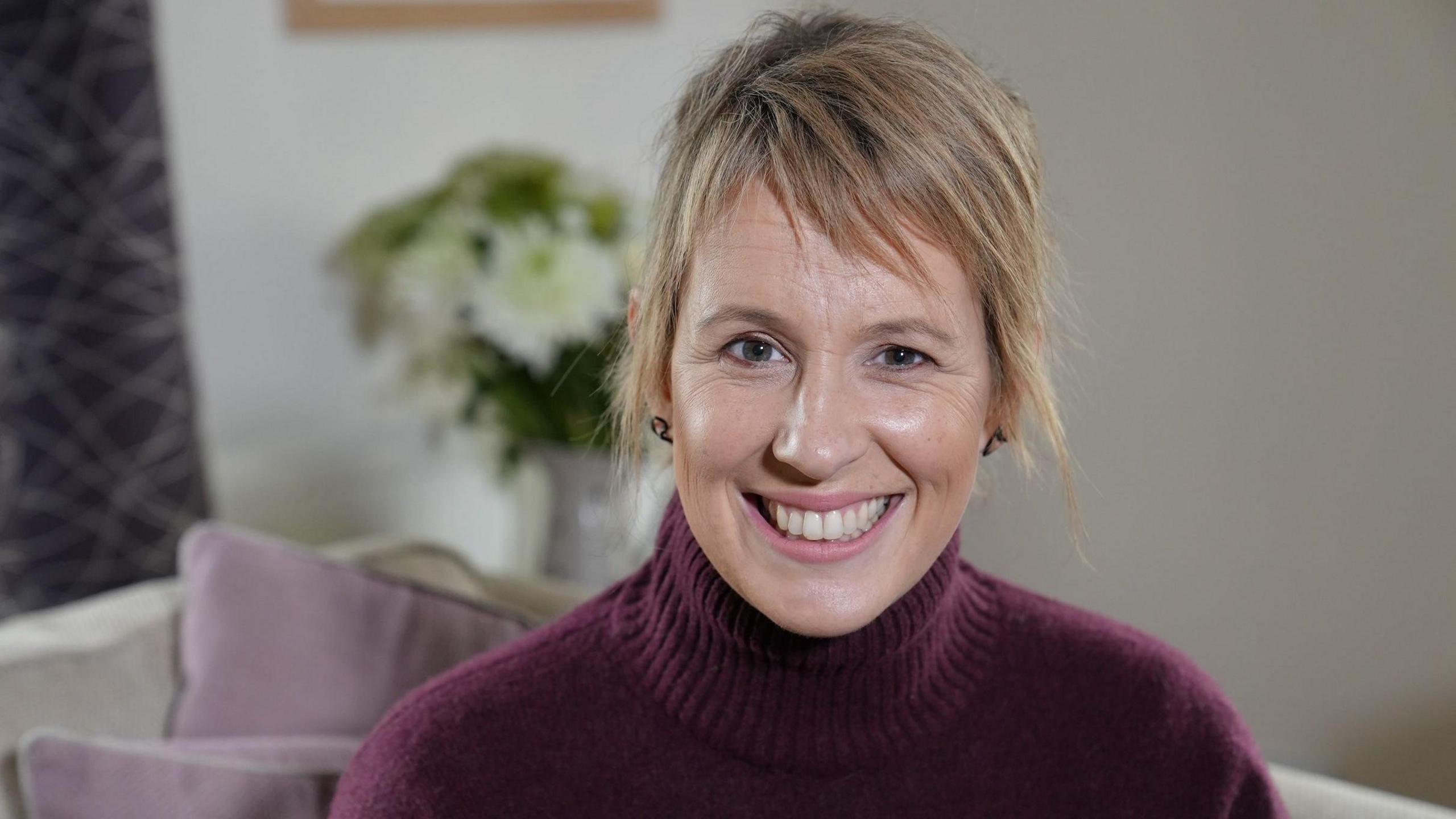 Presenter Mari Grug smiling. She is wearing a purple jumper and her short, dyed blonde hair is tied back. She is sitting on a sofa, which has light-coloured purple cushions.