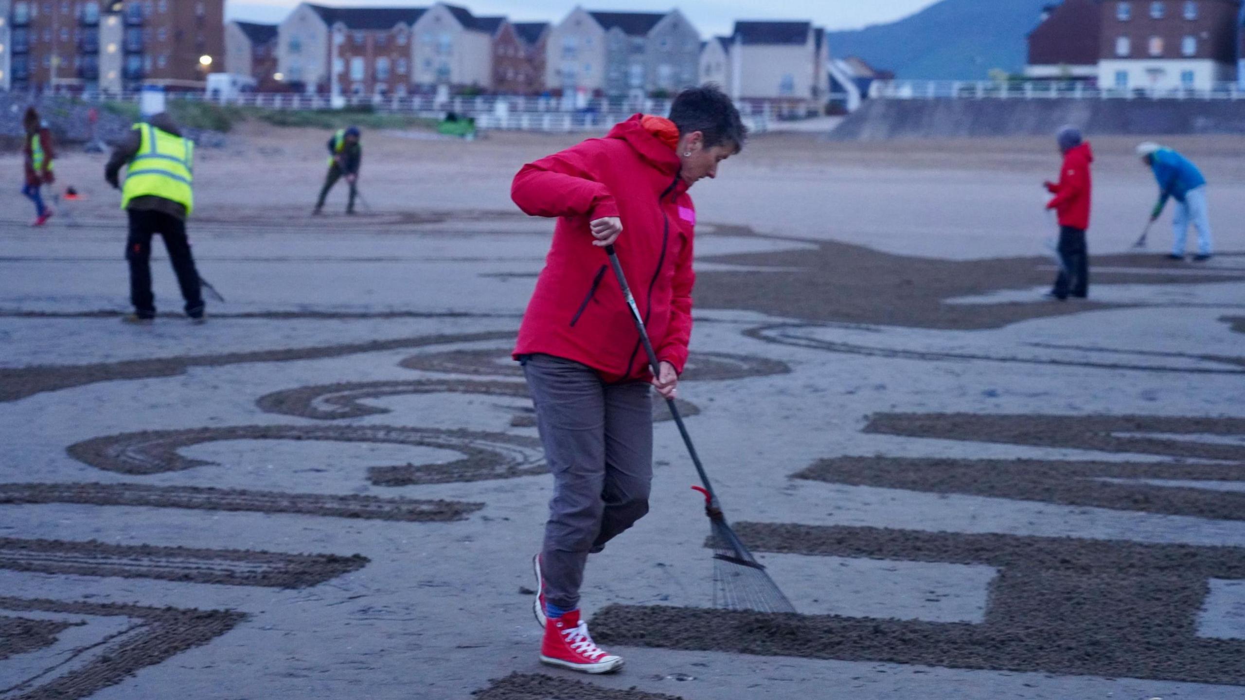 People with rakes etch huge letters into the sand on a beach.