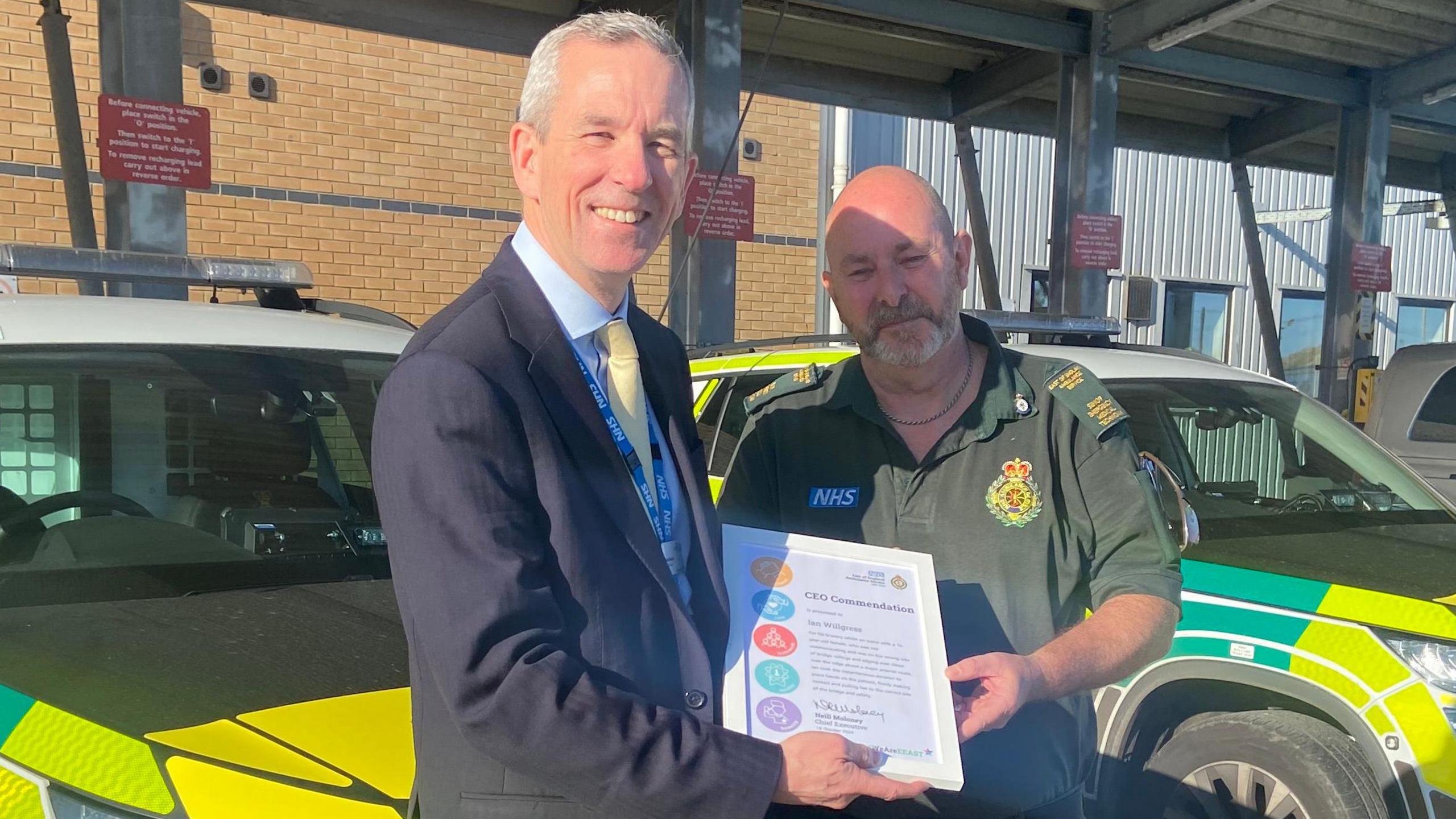 Left, Neill Moloney dressed in a black suit with a light blue shirt and yellow tie is giving Ian Willgress a certification and is facing smiling the camera. On the right is Ian Willgress in green ambulance service uniform and he is looking towards the camera.