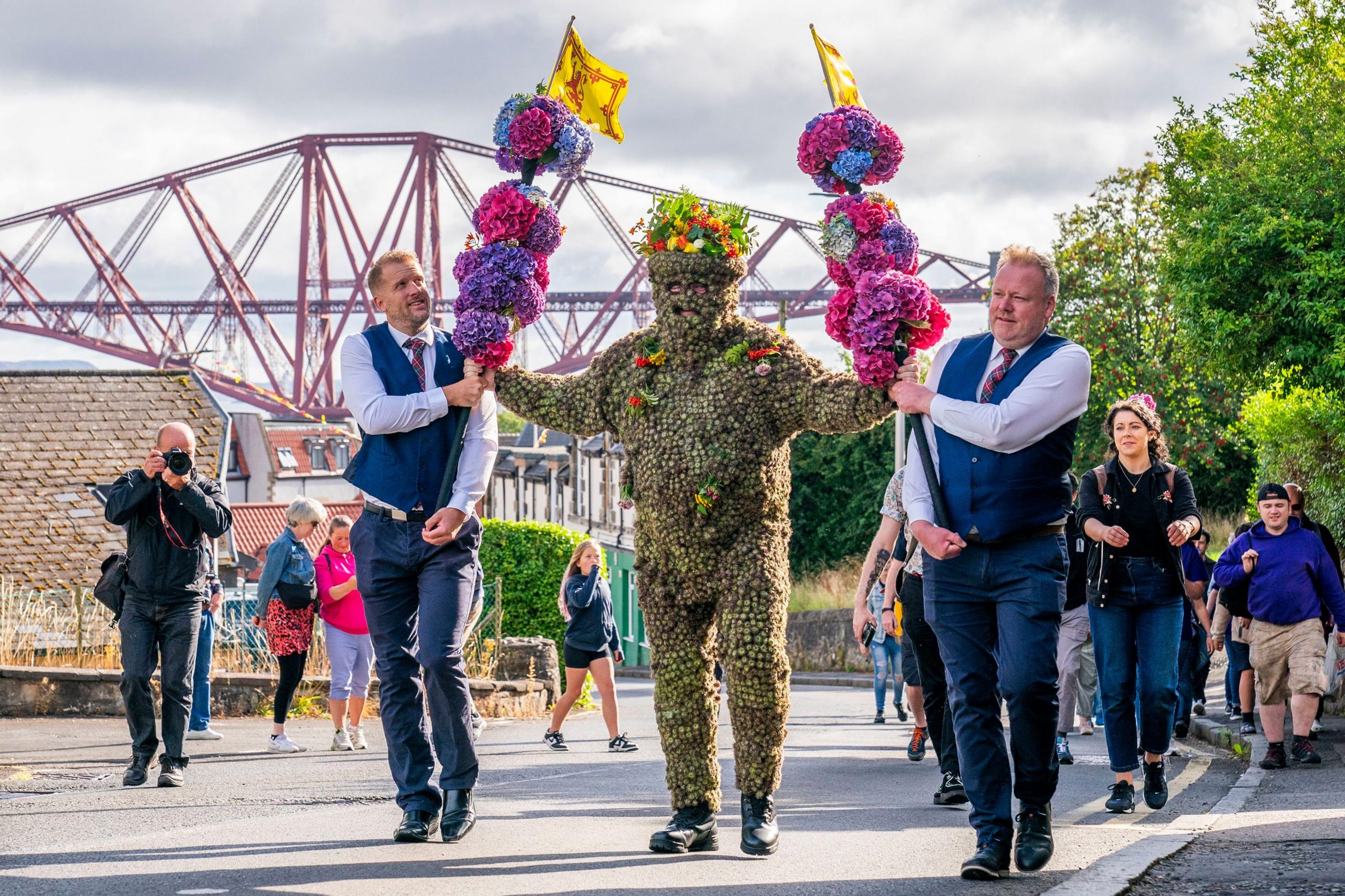 Burryman walking in front of the Queensferry bridge with two minders, helping him carry flower covered staffs