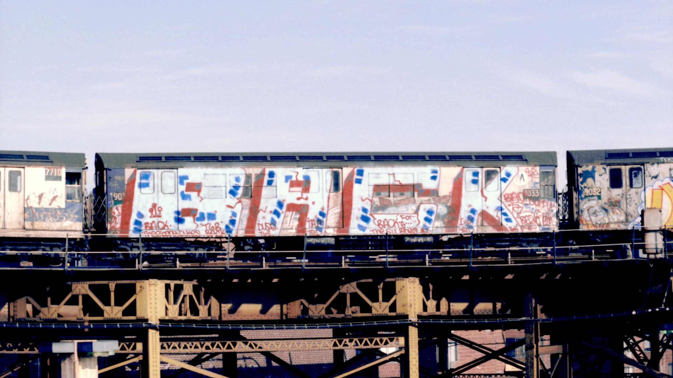 A subway train covered in red, white and blue graffiti on a metal overpass in New York.