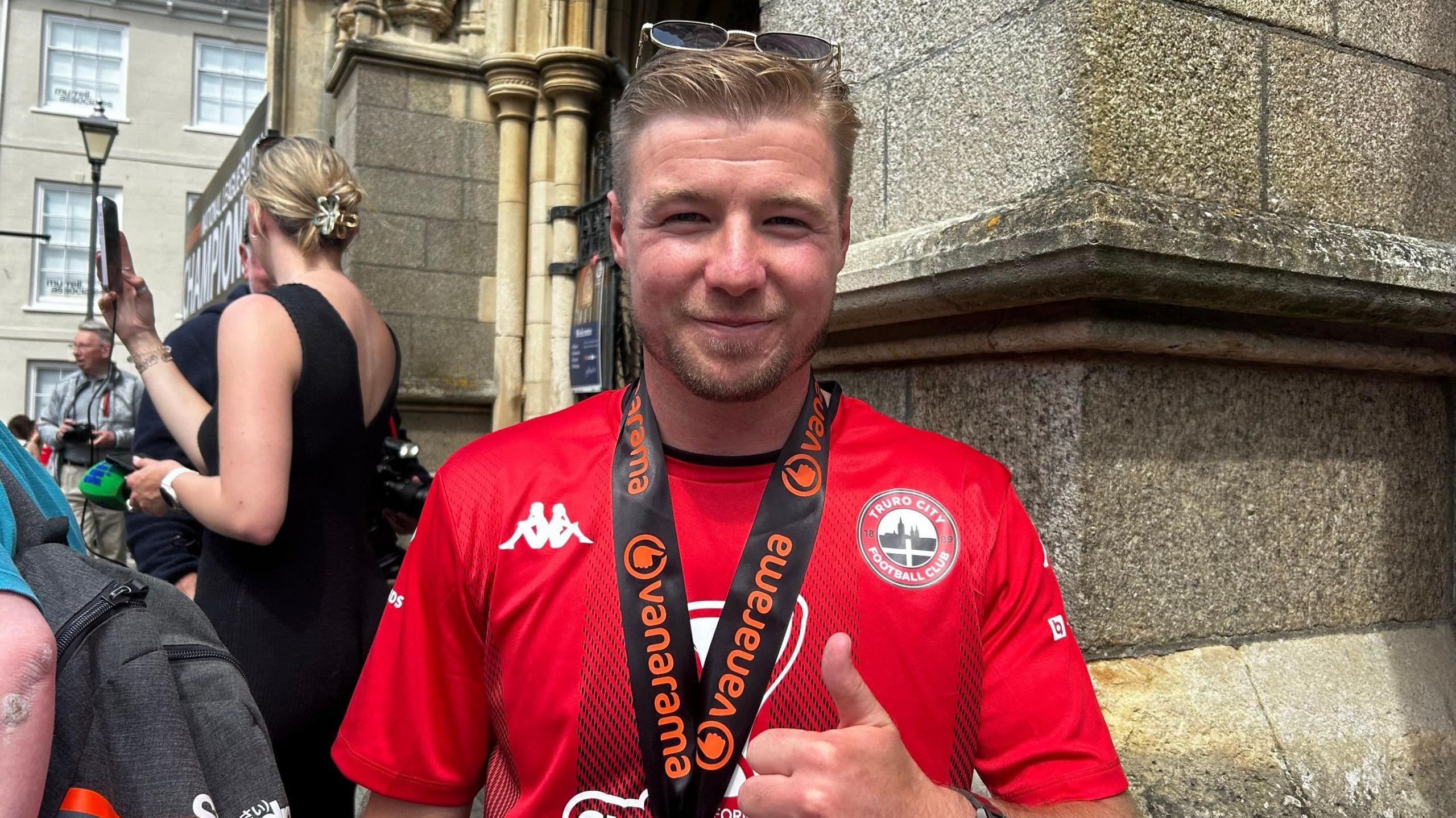 Connor Riley-Lowe is smiling with his thumbs up. He has a medal around his neck and is standing on the steps of the cathedral. There are spectators taking photos behind him.