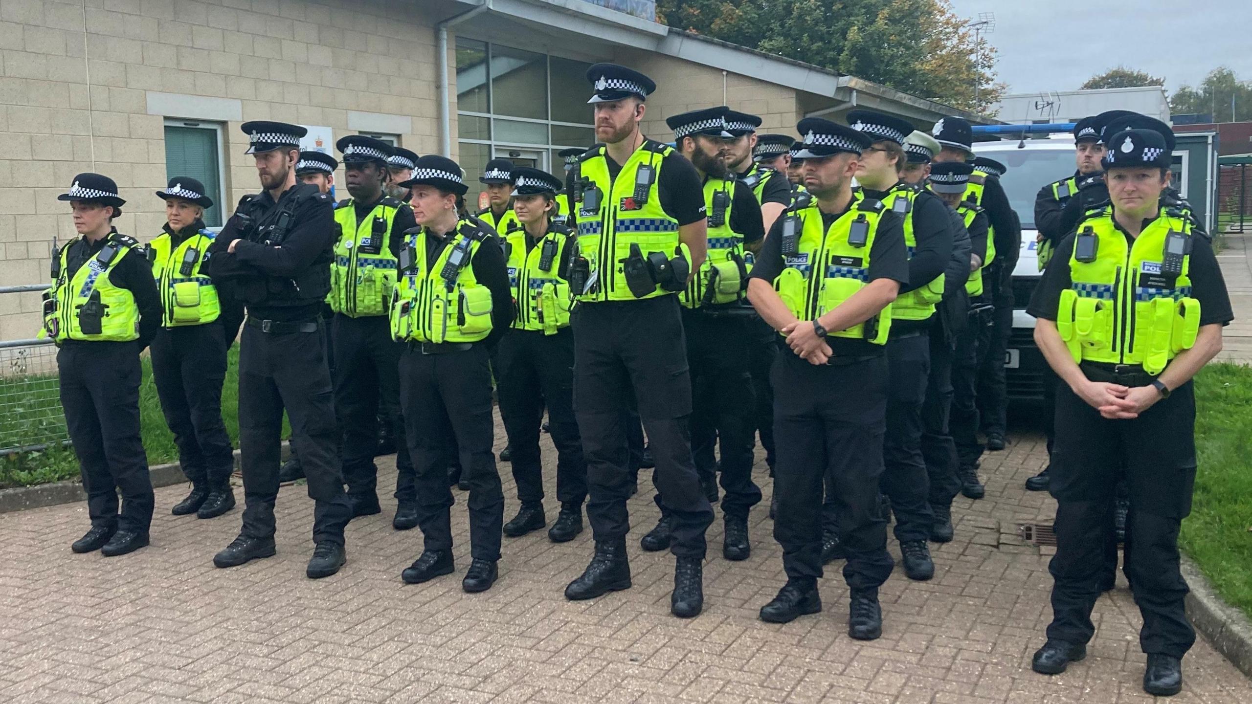 A group of North Yorkshire Police officers stood near a single storey building. All the officers are wearing dark clothing beneath yellow police issue vests complete with body cameras and police radios