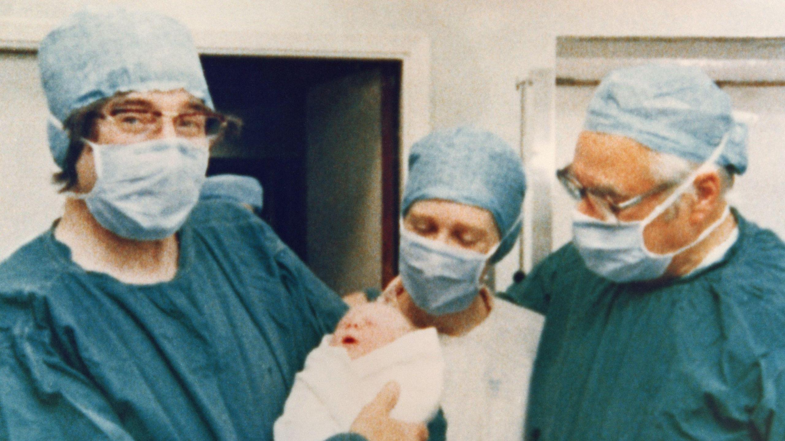 Three medics in green scrubs and surgical masks hold baby Louise shortly after her birth in hospital.