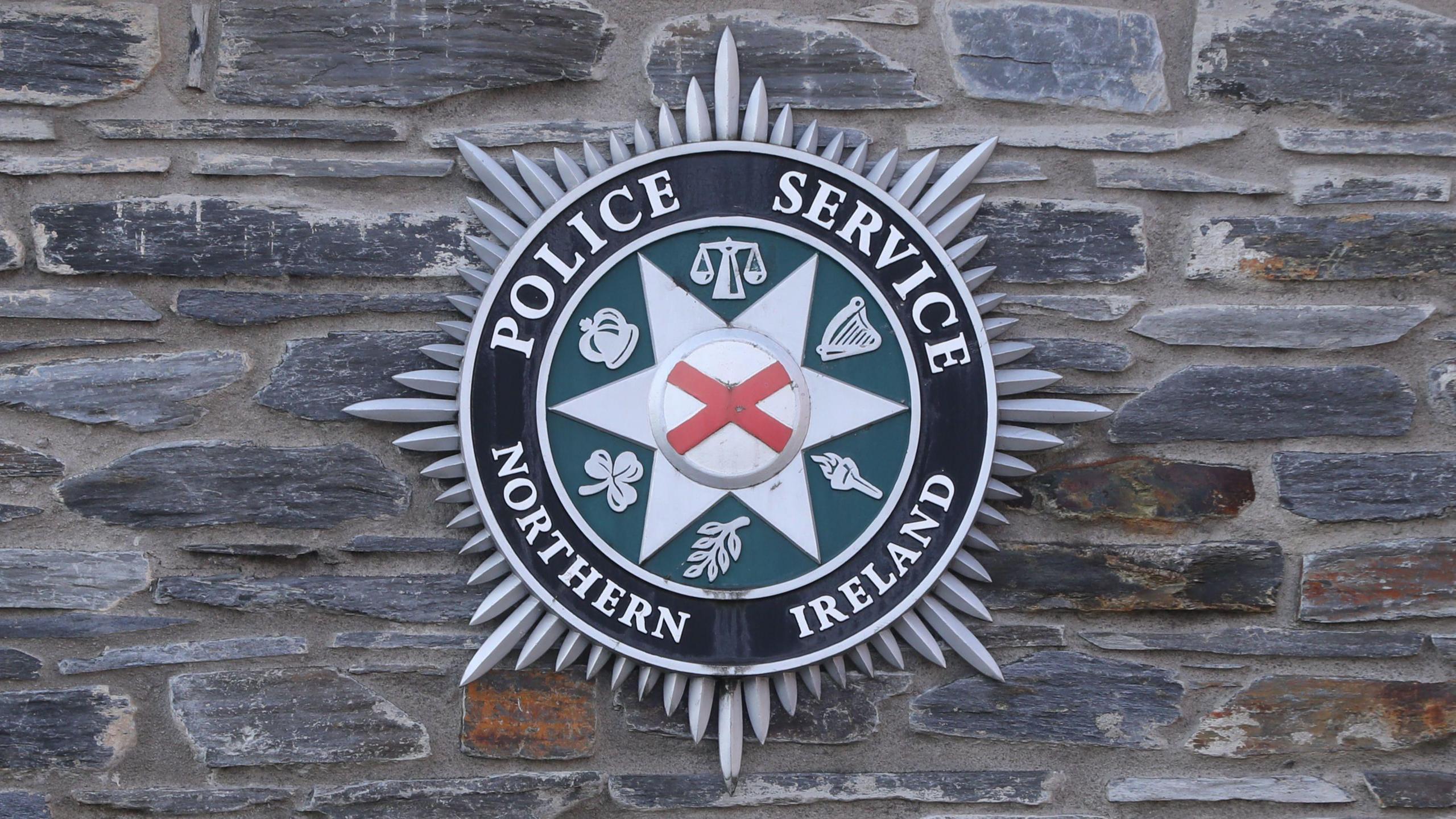 The PSNI logo is on a grey stone wall. The logo has a red ad white cross at its centre, surrounded by six logos on a green background. The words Police service Northern Ireland is in silver letters on a black background. The logo is surrounded by silver spikes. 