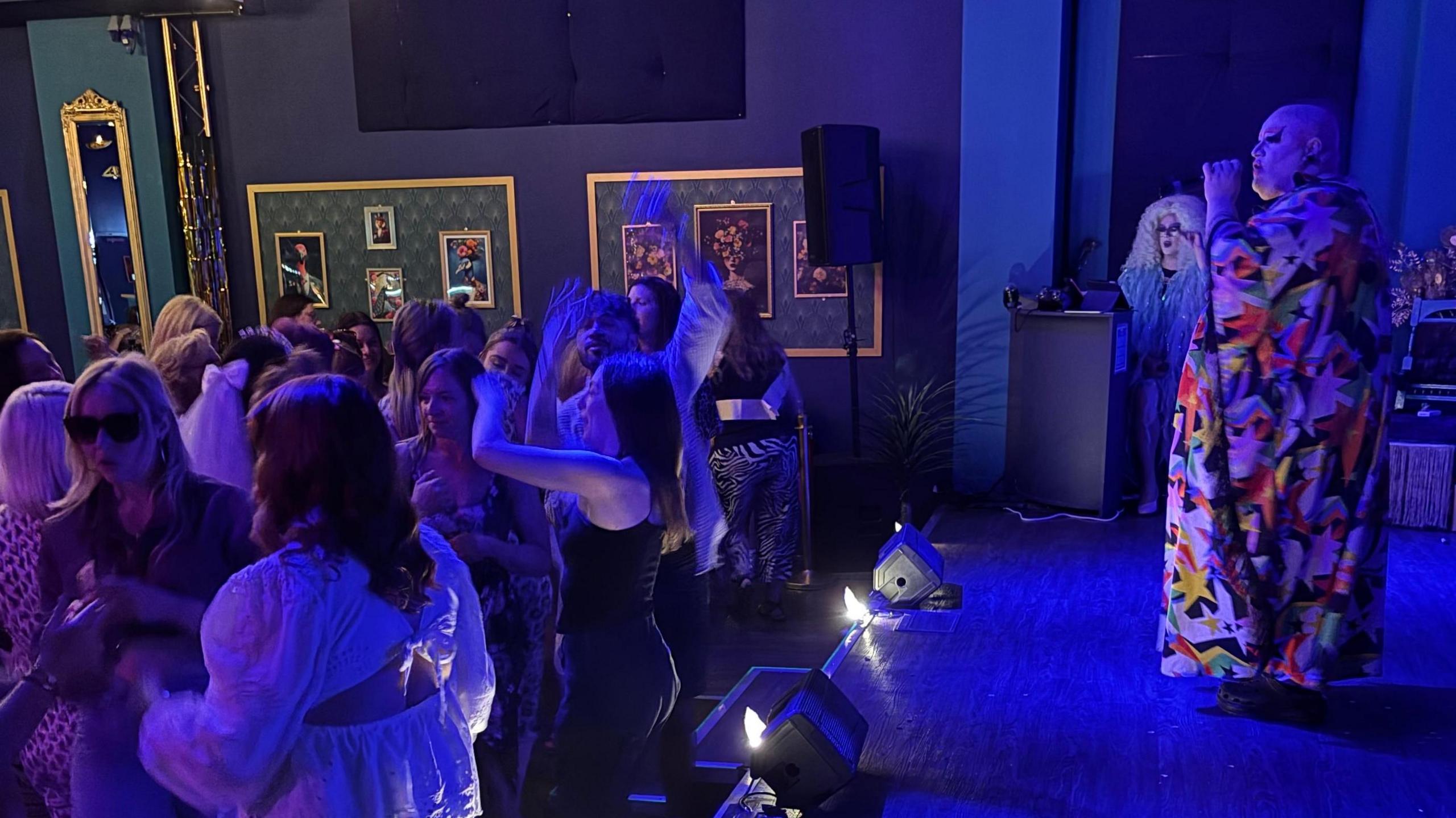 A drag performer is singing on stage in a colourful gown, with people dancing. The room is lit in blue.