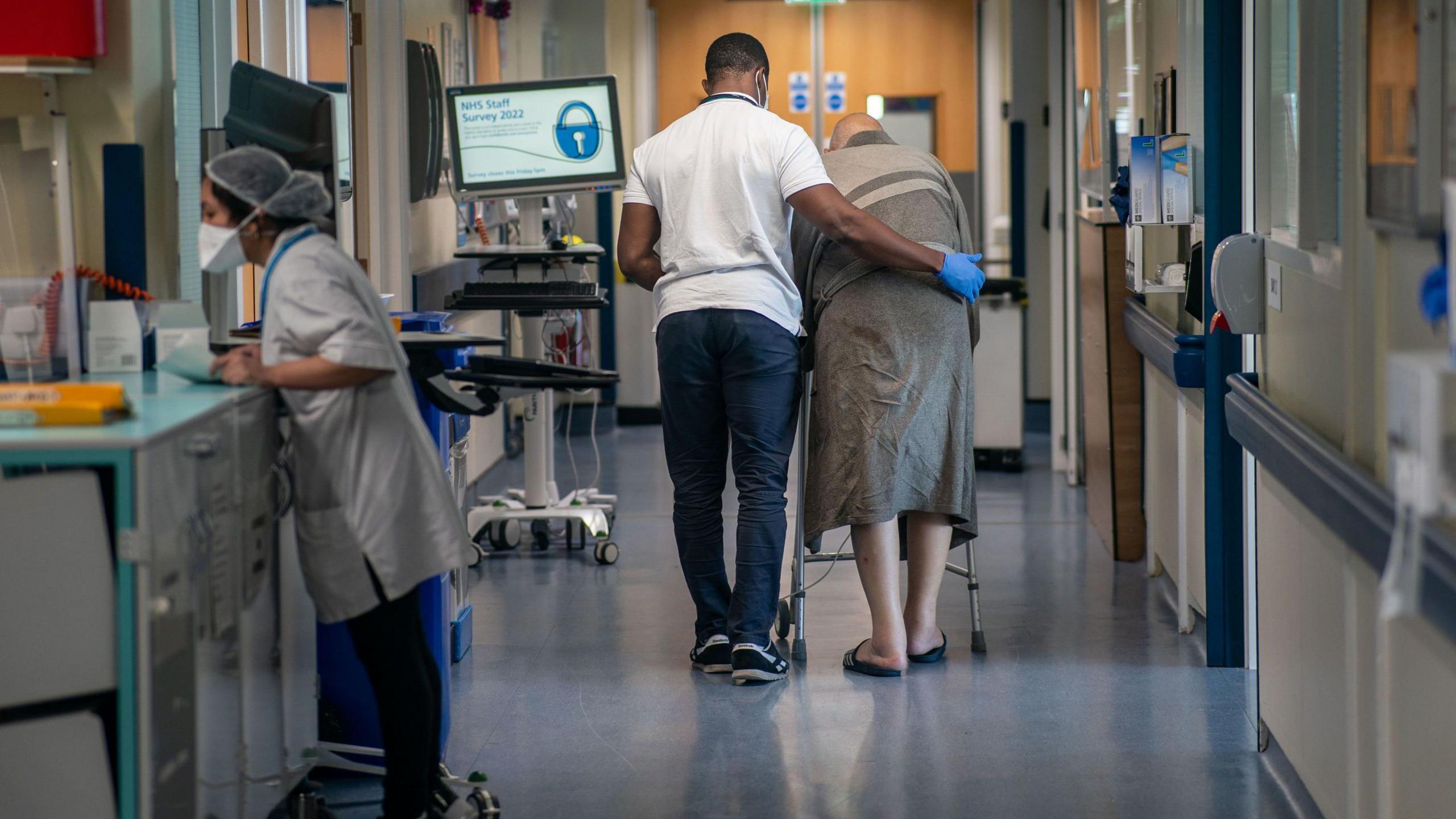 Nursing auxiliary helps an elderly patient with a zimmer frame to walk down a hospital ward