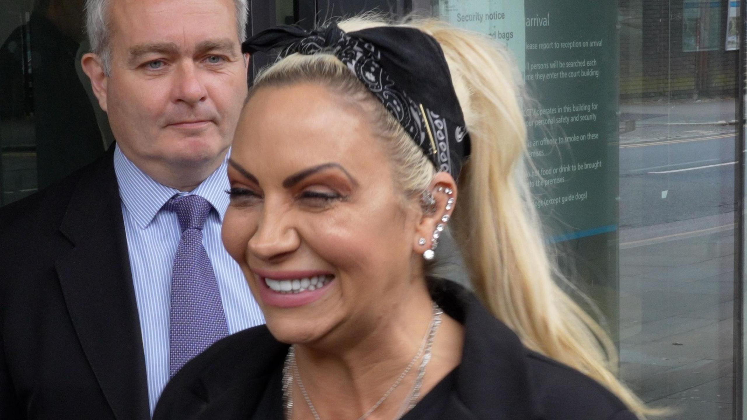 Jodie Marsh squinting while giving a big smile. She has long blonde hair in a ponytail, held in place by a black hair tie. She is wearing black in front of her lawyer, who is wearing a suit.