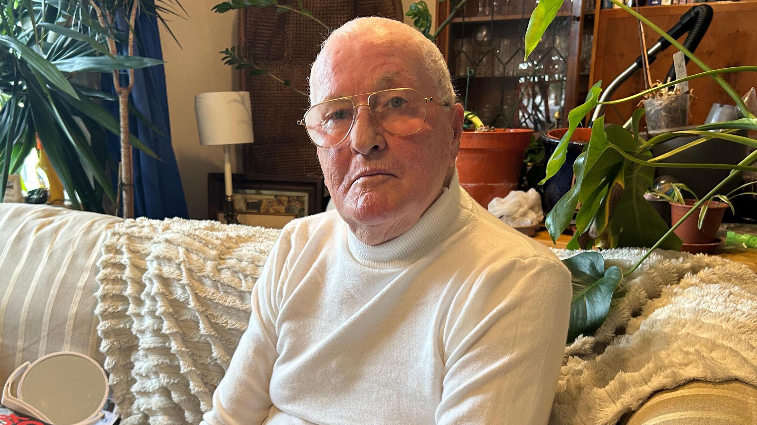 Timothy Willis is pictured sitting on a striped sofa which is cream in colour. There is a fluffy cream blanket draped over the sofa and a sideboard and plants behind it. Mr Willis, who is an older man, is wearing a cream roll-neck jumper and glasses and he has white hair. He has a serious expression and is looking directly at the camera.