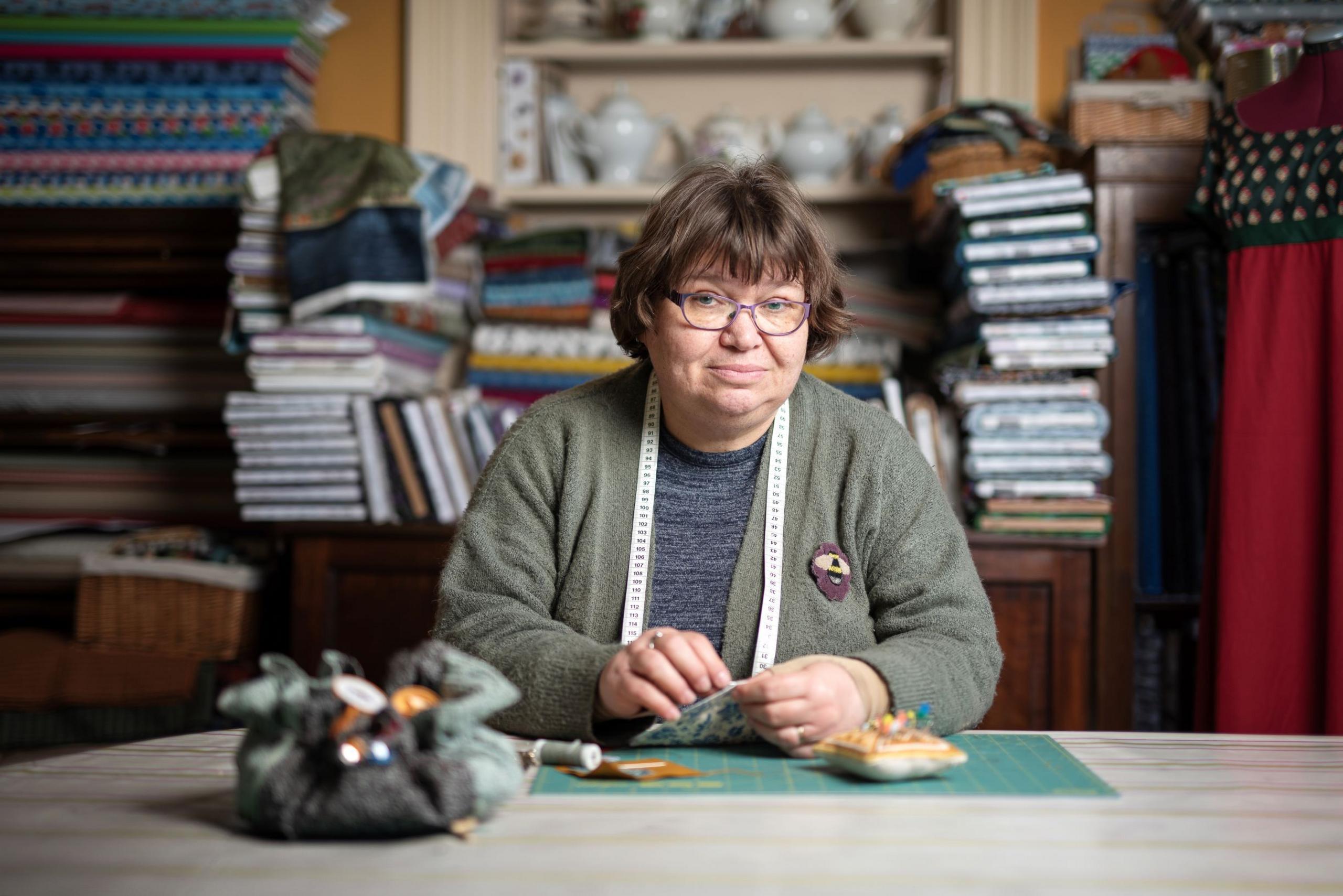 Sad woman with glasses and measuring tape round neck sews at a table, surrounded by piles of books, and looking at camera.