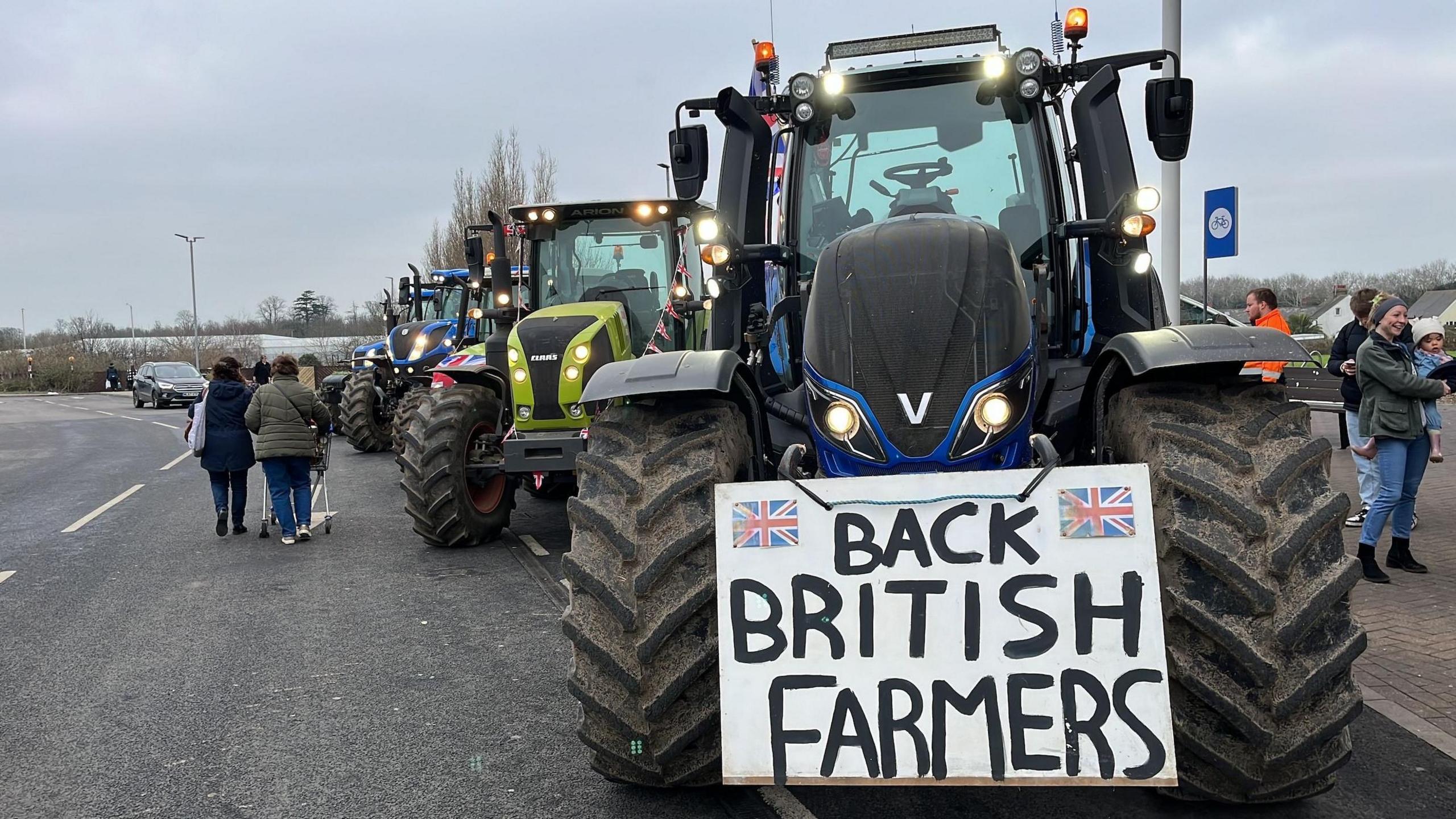 A blue tractor is parked in a supermarket car park and has a sign on its hood reading: "Back British farmers." Behind is a line of parked tractors.