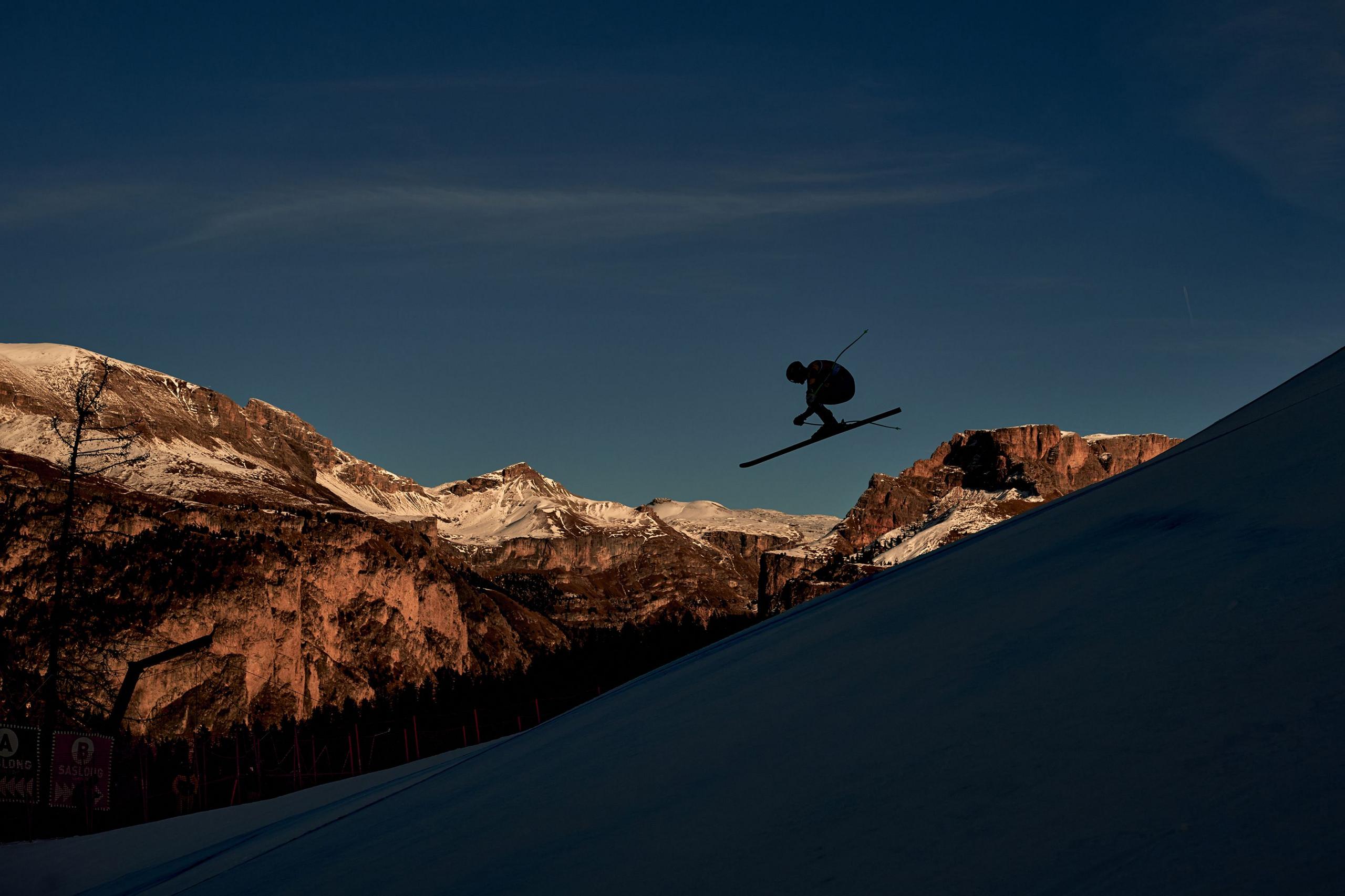A silhouetted skier performs an aerial trick against a deep blue evening sky, captured mid‑jump above a snowy slope. Rugged, sunlit mountain peaks frame the background, glowing in warm tones as the skier’s form contrasts sharply with the darkening landscape.
