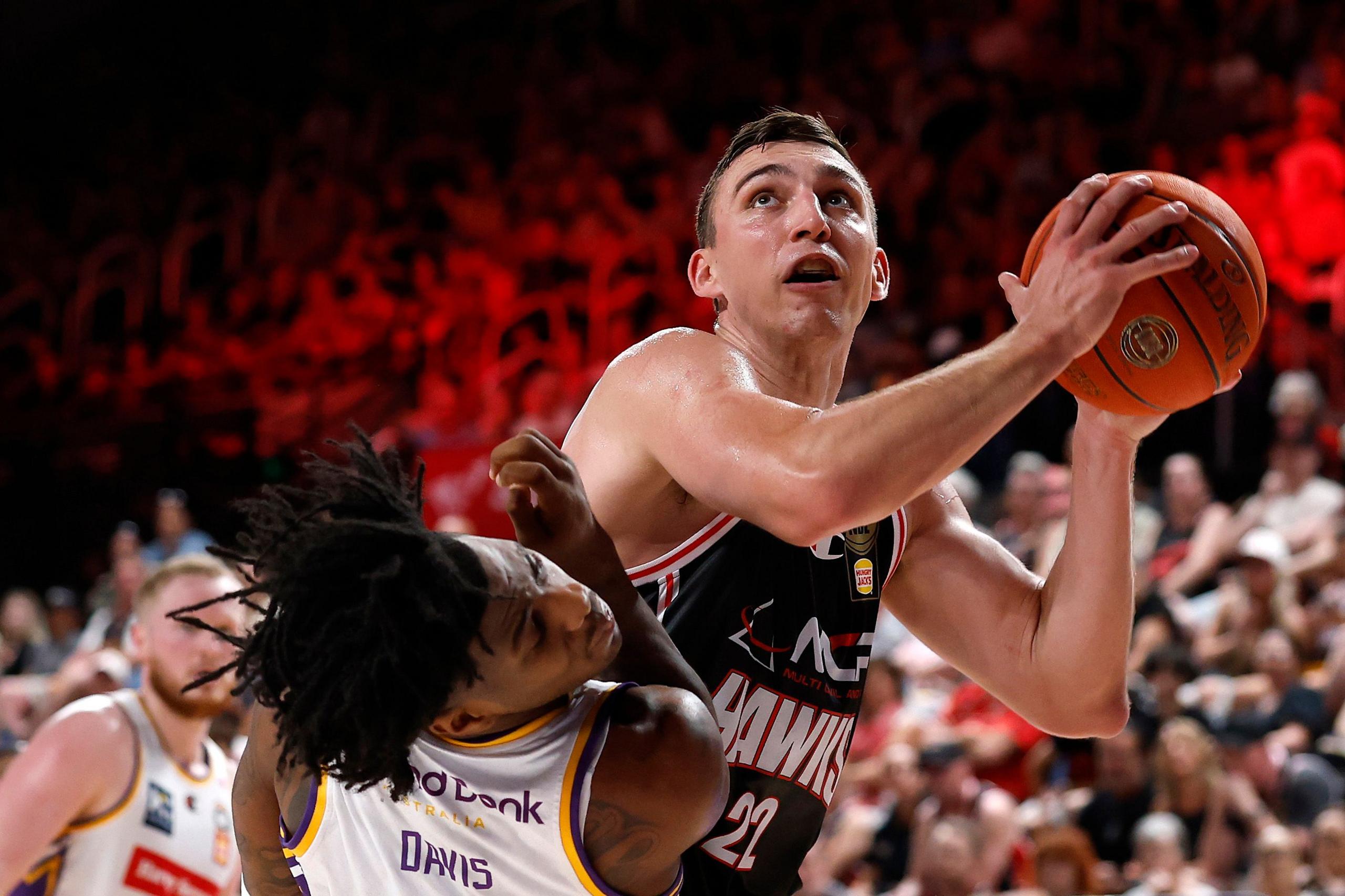 Basketball player in a black jersey holding the ball close to the hoop while an opposing player in a white jersey attempts to block, with a packed arena and red lighting in the background.