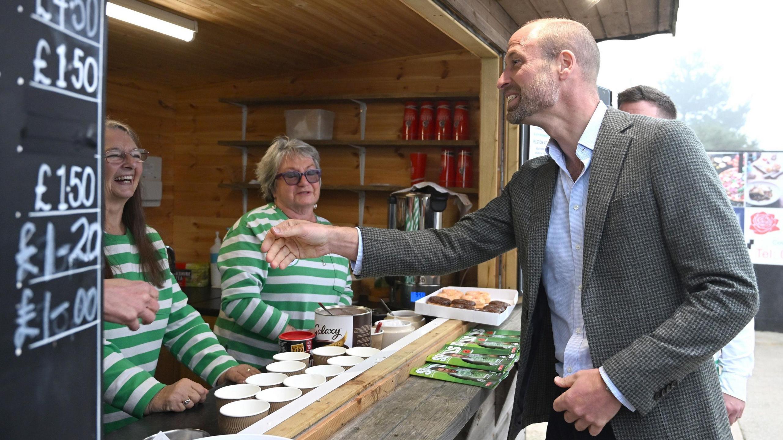 Prince William is laughing as he reaches to shake a woman's hand. Three women are manning a hut which sells teas, coffees and cakes. They are wearing green and white striped football tops.