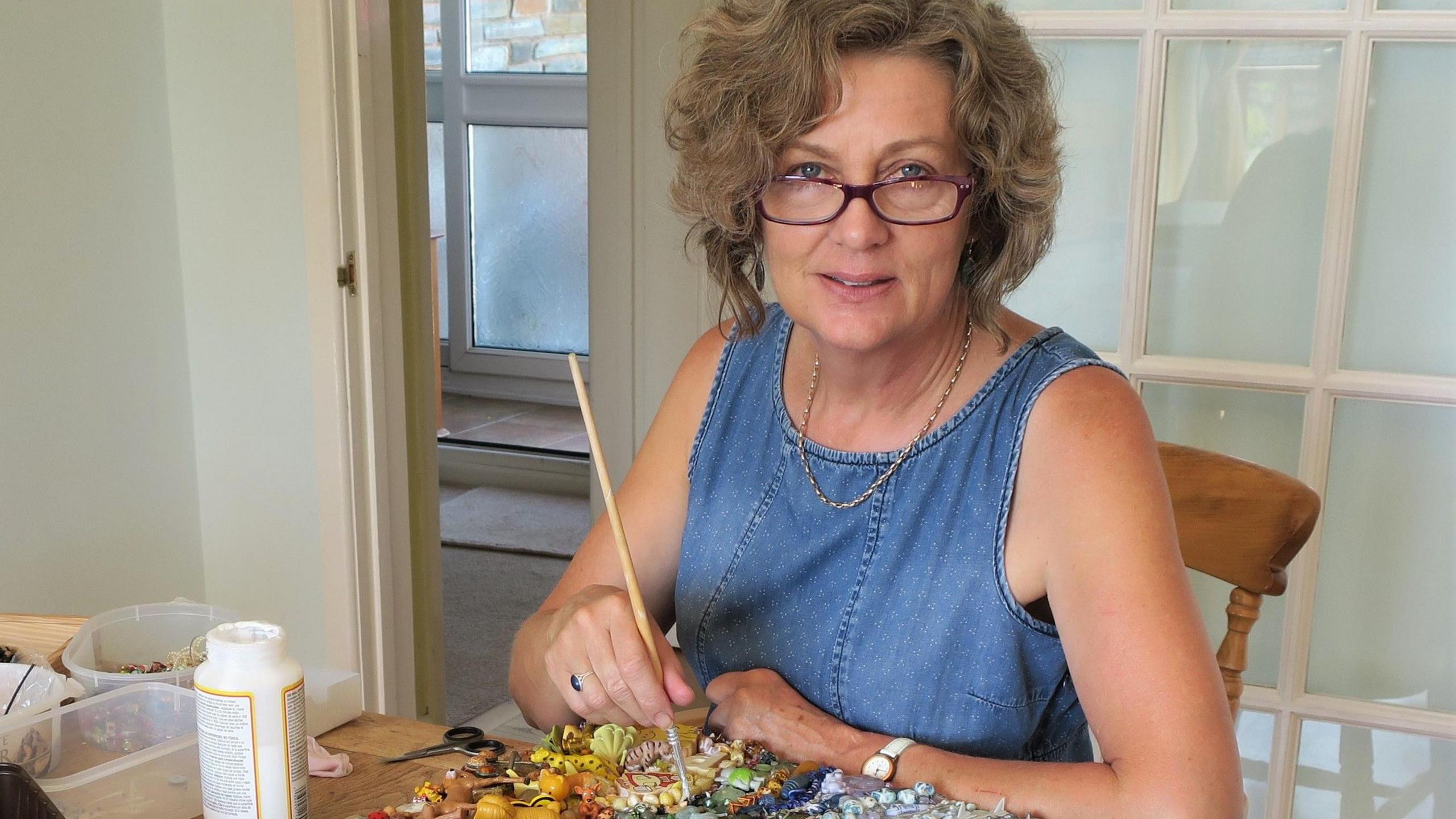 An artist holds a paintbrush over a collage of tiny plastic items and a pot of glue. She's making a brightly coloured piece inspired by the painter van Gogh