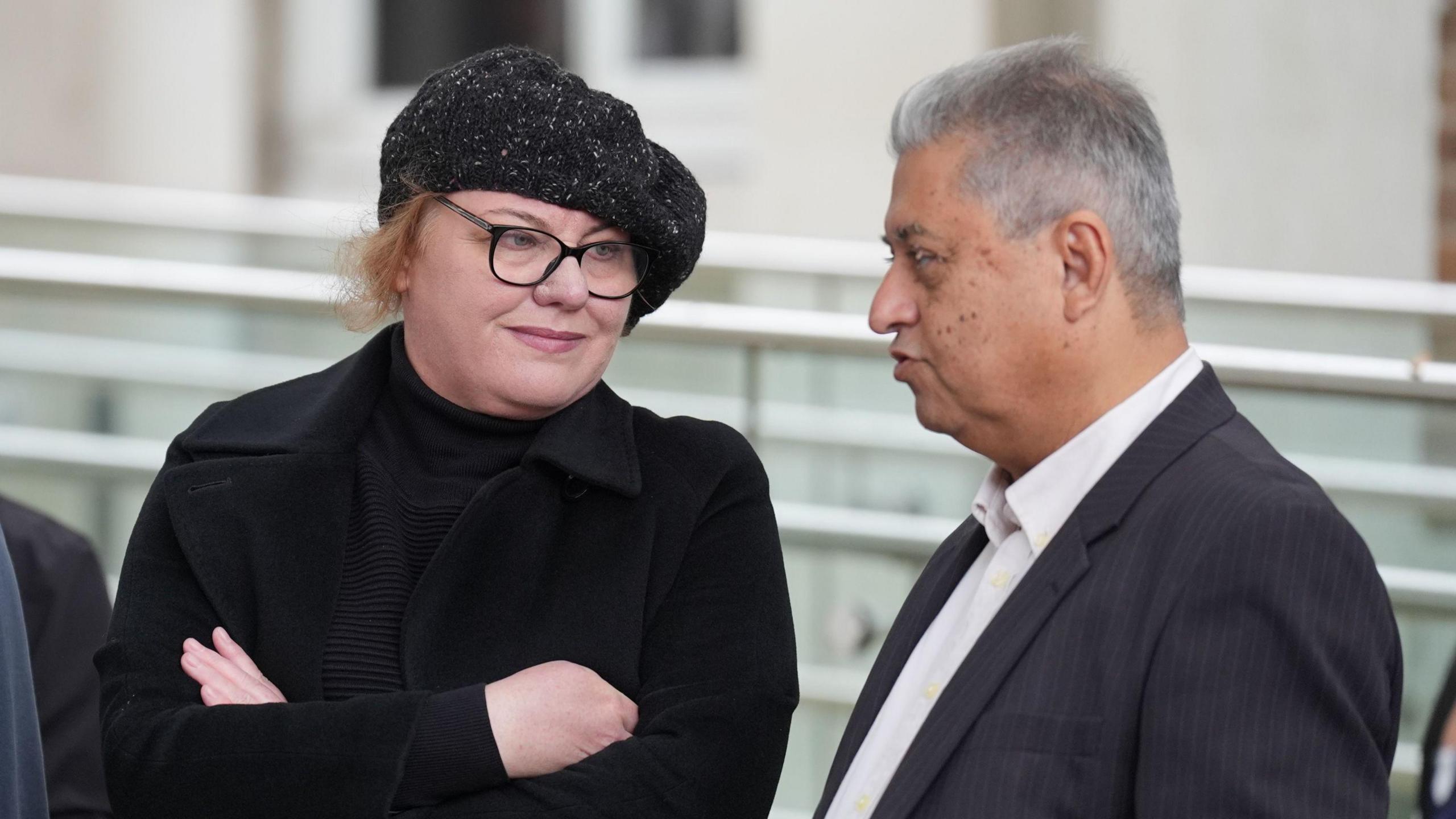 Julia Hopper talks to Kobad Bankwala, 68, the father of Darian Bankwala who was 22 years old when he was hit by a train and killed near Wickford station in Essex on December 27 2020, outside the Lampard Inquiry at Chelmsford Civic Centre. Ms Hopper  is wearing a black coat and black polo neck jumper, black glasses, and a black hat. Mr Bankwala is wearing a grey suit and white shirt.