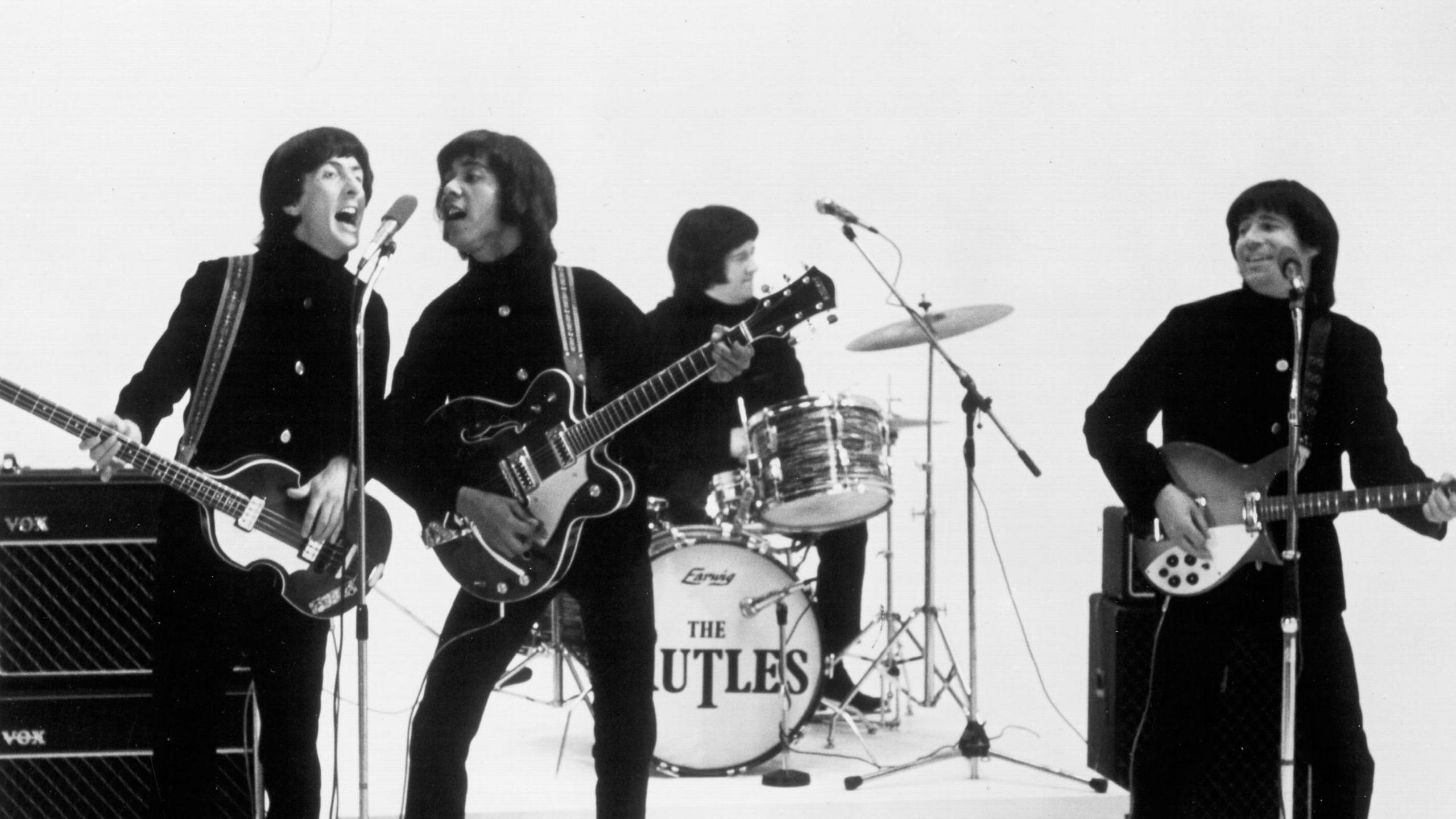 Black and white image of The Rutles (left to right - Eric Idle as Dirk and Ricky Fataar as Stig sing into the same microphone as they play their guitars, with John Halsey as Barry playing a drum kit branded with The Rutles and Neil Innes as Ron playing a guitar in front of a microphone stand).