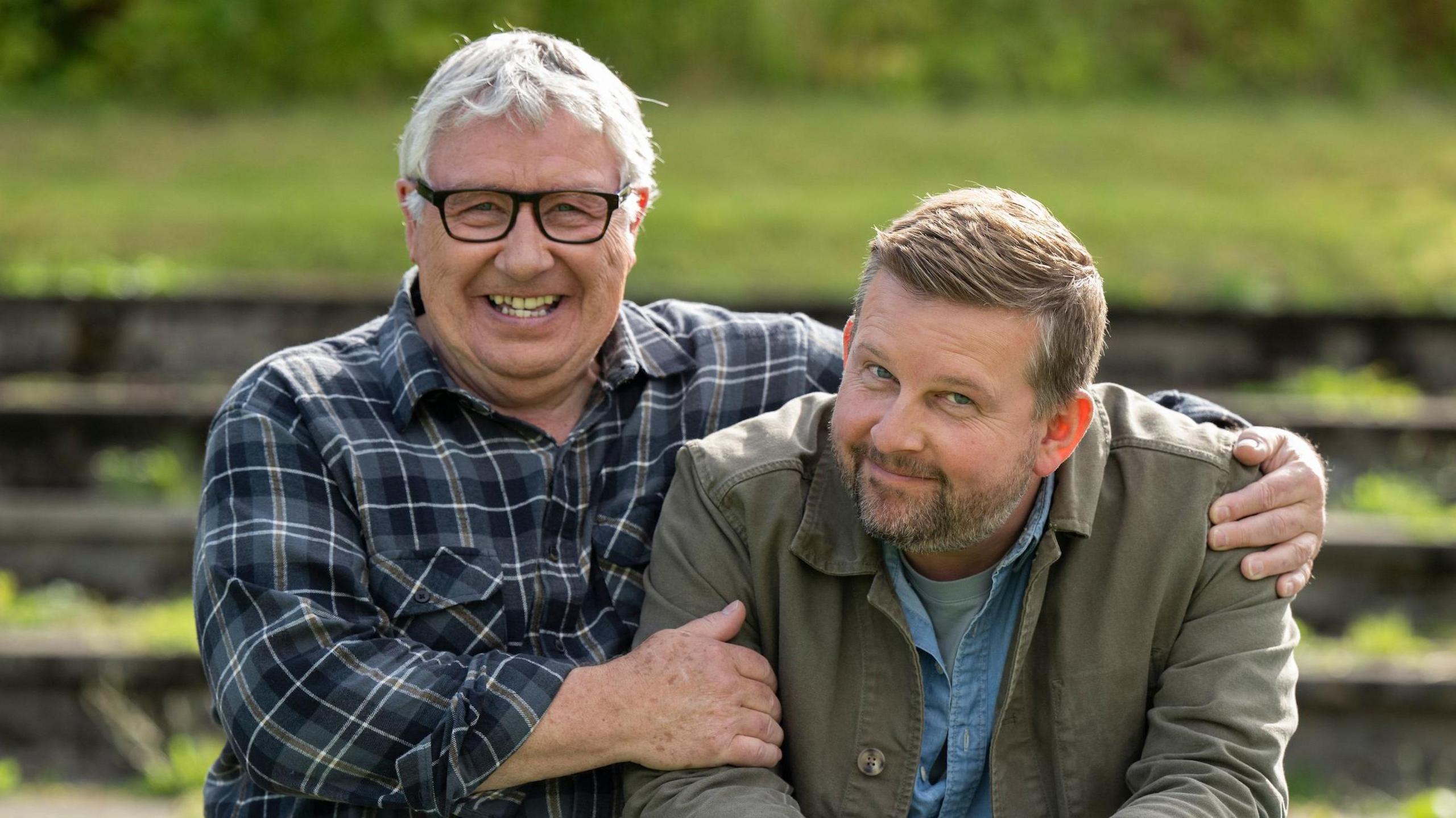 A bespectacled man with grey hair sits with his arms around the shoulders of a younger bearded man