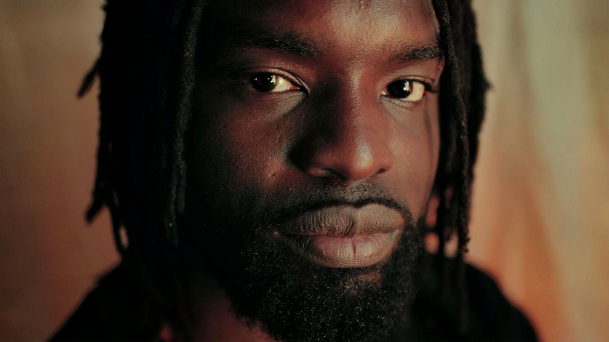 A close-up of Meji Alabi, with beard and dreads, looking into the camera