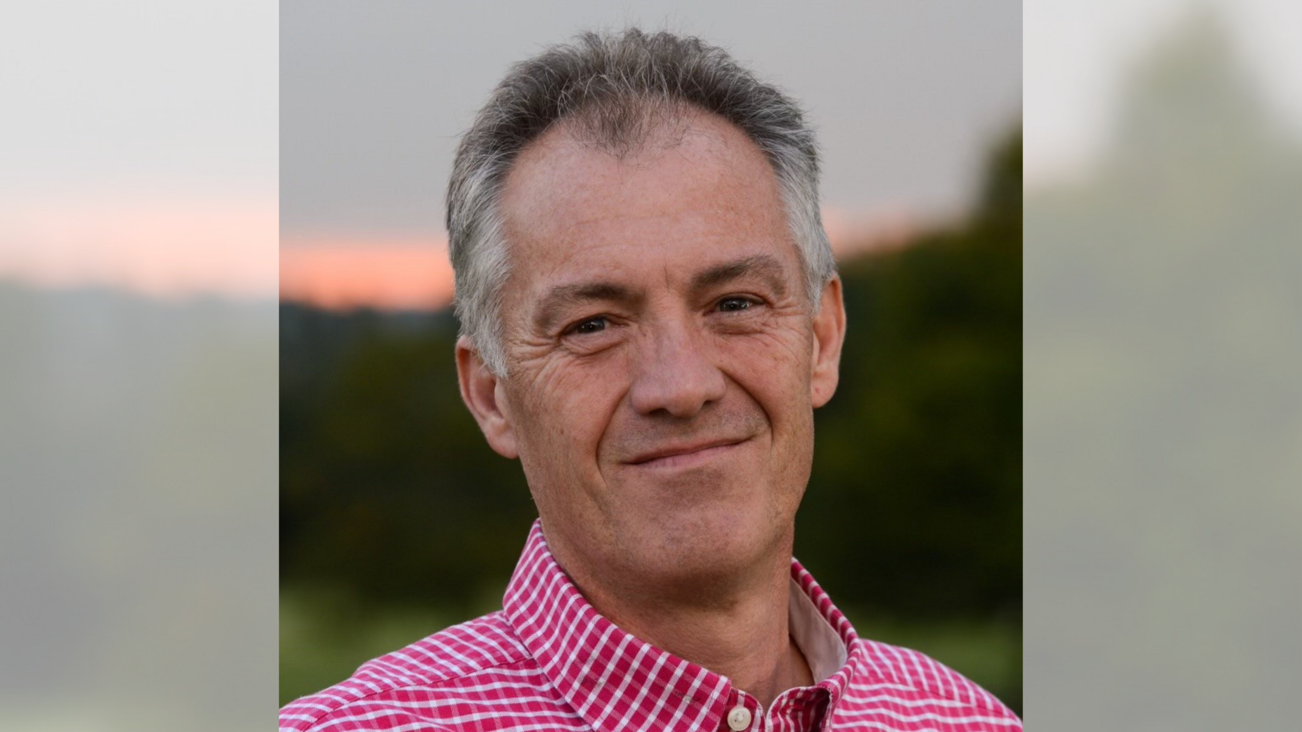 A man with a red and white check shirt smiling at the camera in front of a blurred sunset