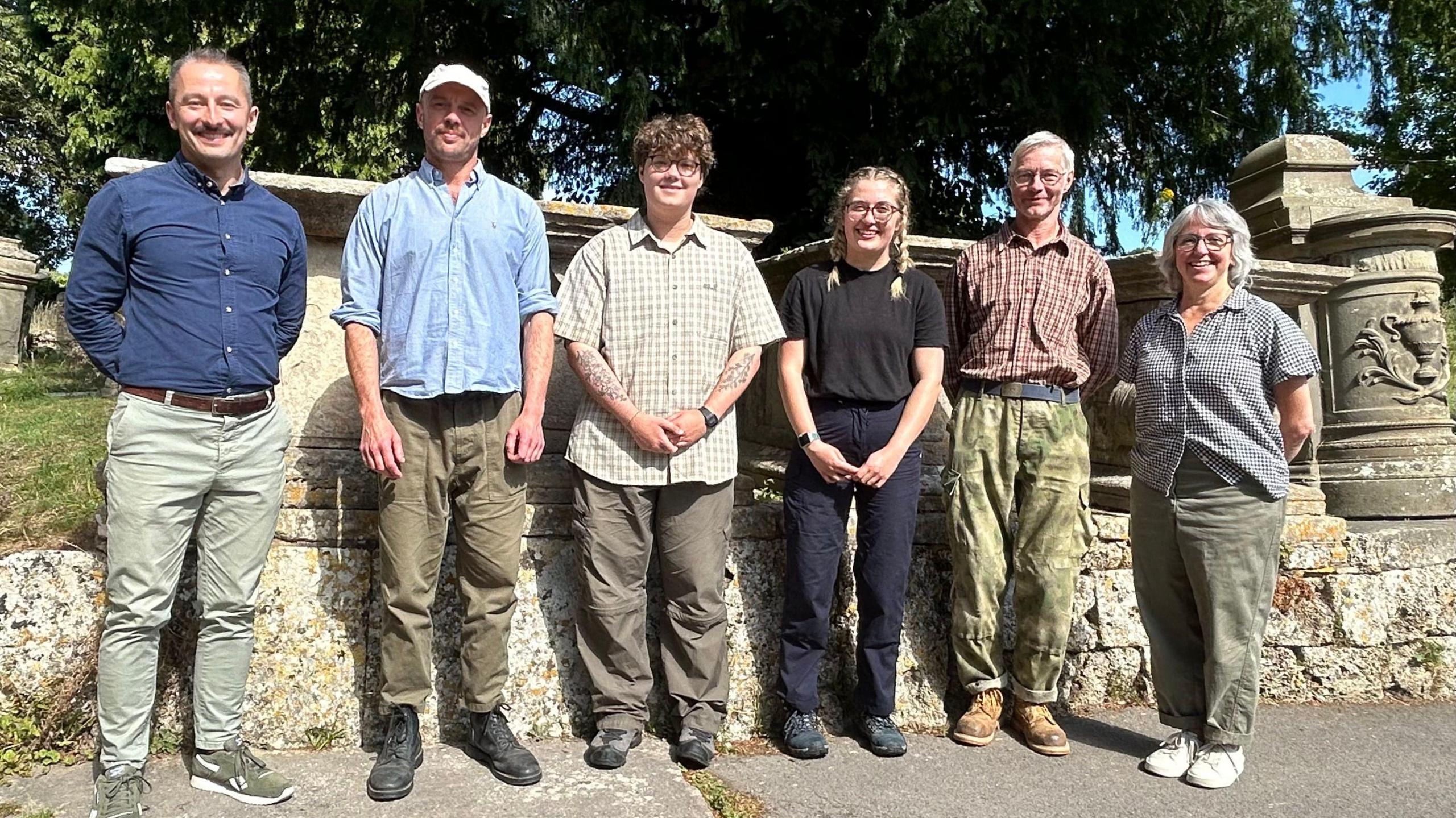 SIx people made up of the project team with conservation students, standing in front of some of the monuments and all smiling at the camera. 
Behind them and on the left half you can see the flat top that is distinctive of these stones. 
There are trees and grass behind them. 