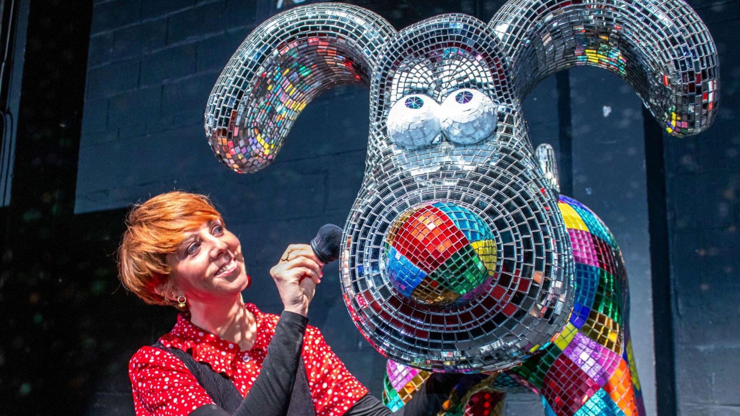 A woman with ginger hair polishes a Gromit statue at the launch of the Gromit Unleashed 3 trail in Bristol. The statue is covered in individual pieces of sparkling material, and is in many colours. The woman is wearing a red and white shirt with a black dress