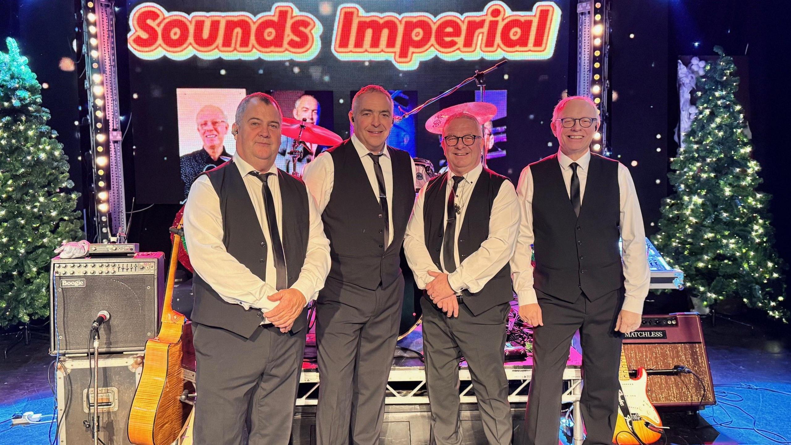 Four men wearing black suits and white shirts standing in front of a small stage and a sign saying Sound Imperial