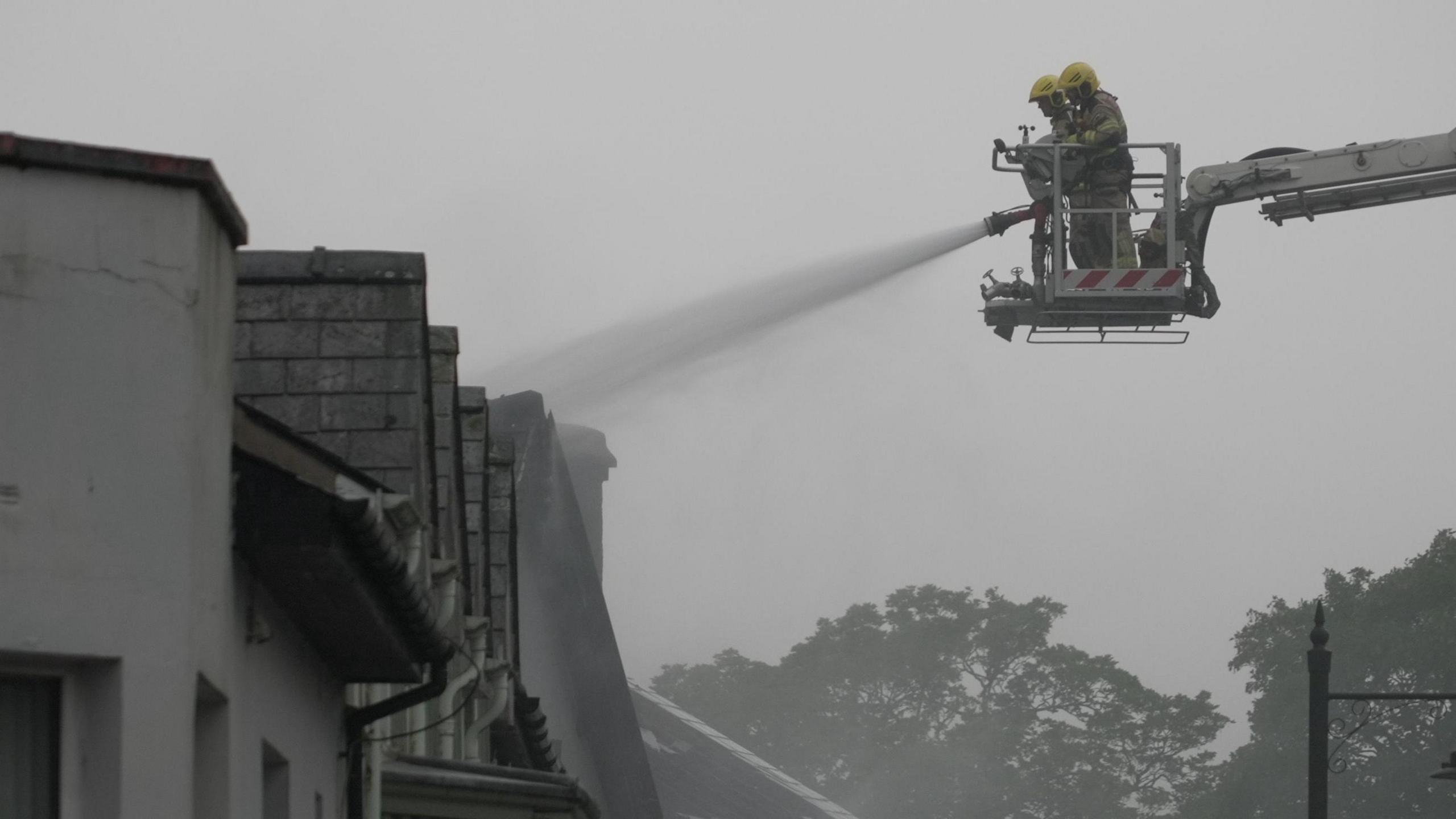 Two fireman in a platform on a crane firing a large amount of water from a jet hose on to a building. There is a film of fog or smoke over the building