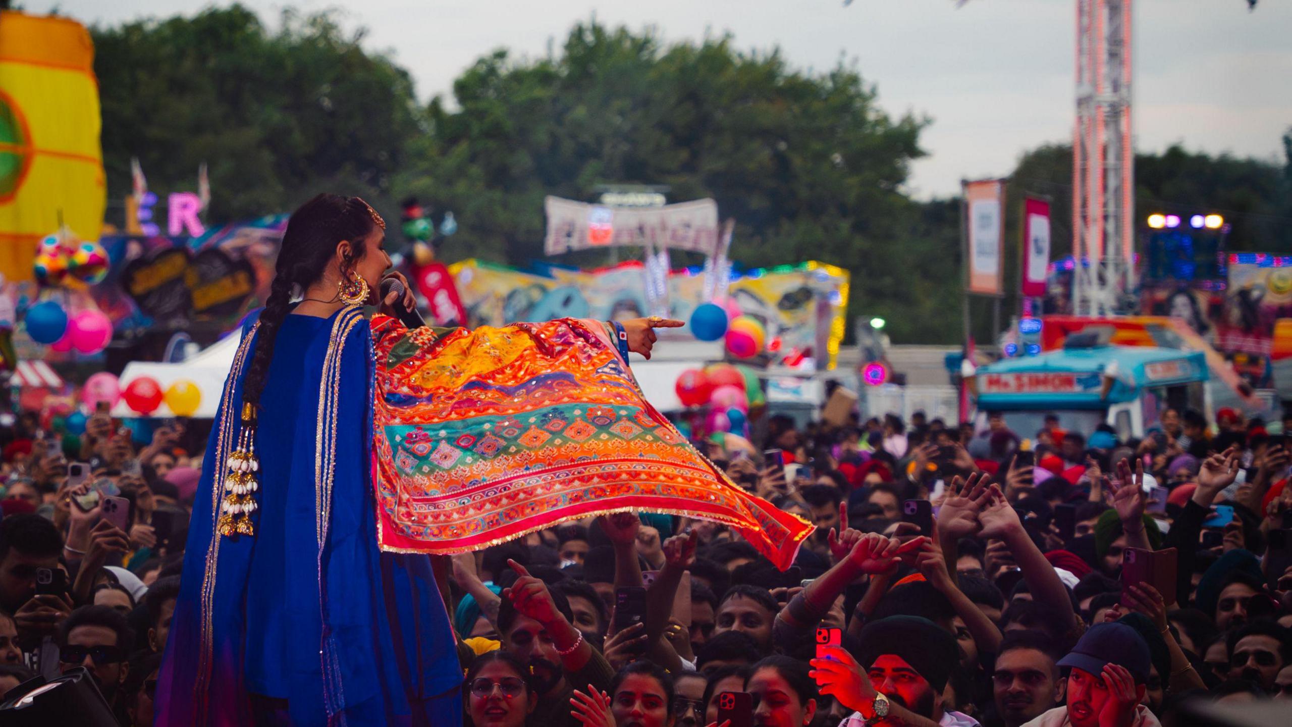 A woman speaks into a microphone and points as she stands on a stage in front of a crowd of people. A fairground is visible in the distance behind the crowd.