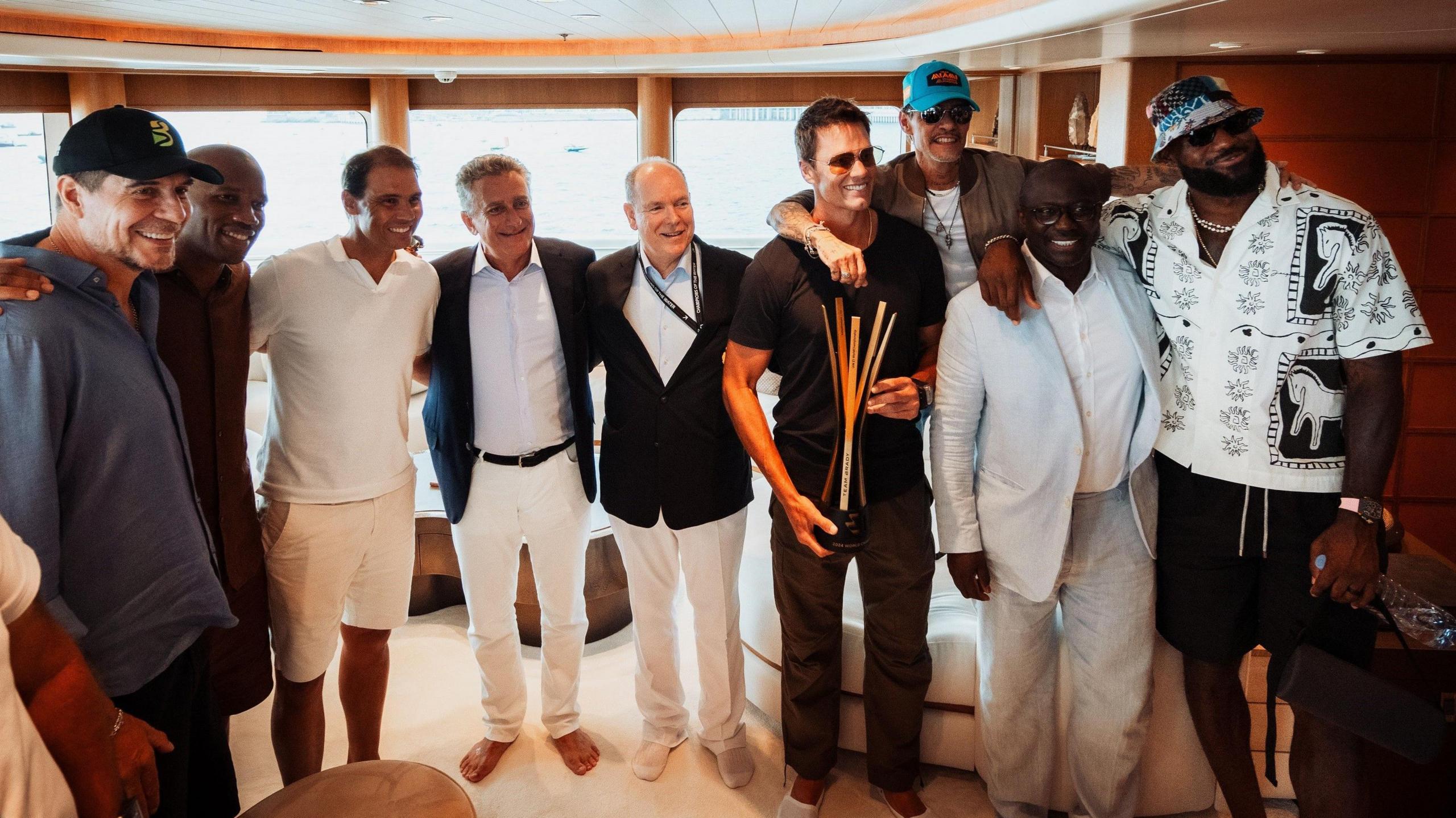 A group of nine men including Marcelo Claure, Didier Drogba, Rafael Nadal, Prince Albert II of Monaco, Tom Brady, Marc Anthony and LeBron James pose with their arms around each other for a photo. They are standing in the plush interior of a boat with wood veneer and soft furnishing, while Tom Brady is holding a tall trophy made of several individual vertical pieces of metal
