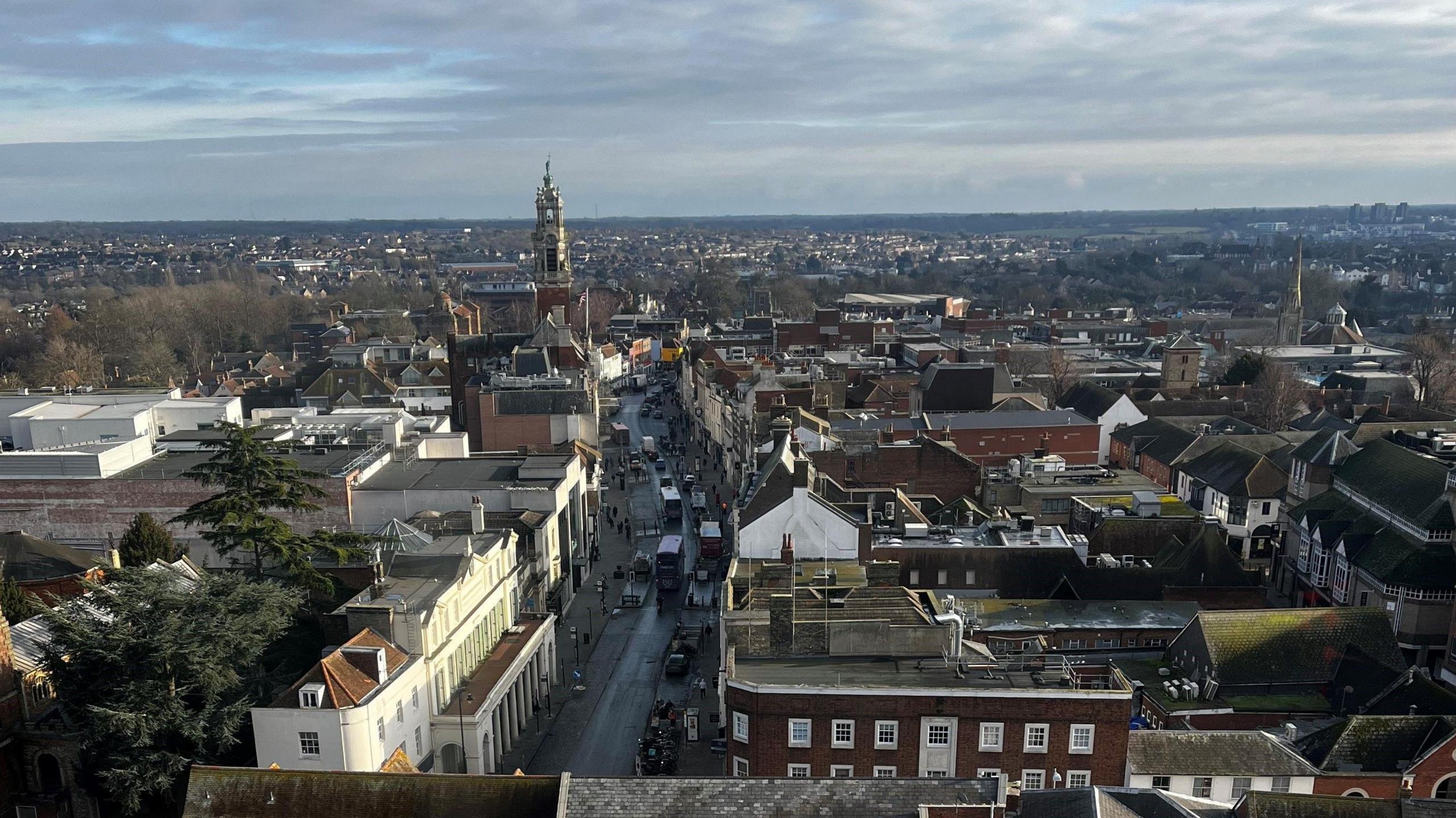 An aerial view of Colchester city centre. It shows the high street, with shops either side. On the left, about halfway down, the Italianate town hall's tower rises above the city. The view stretches to countryside on the horizon.