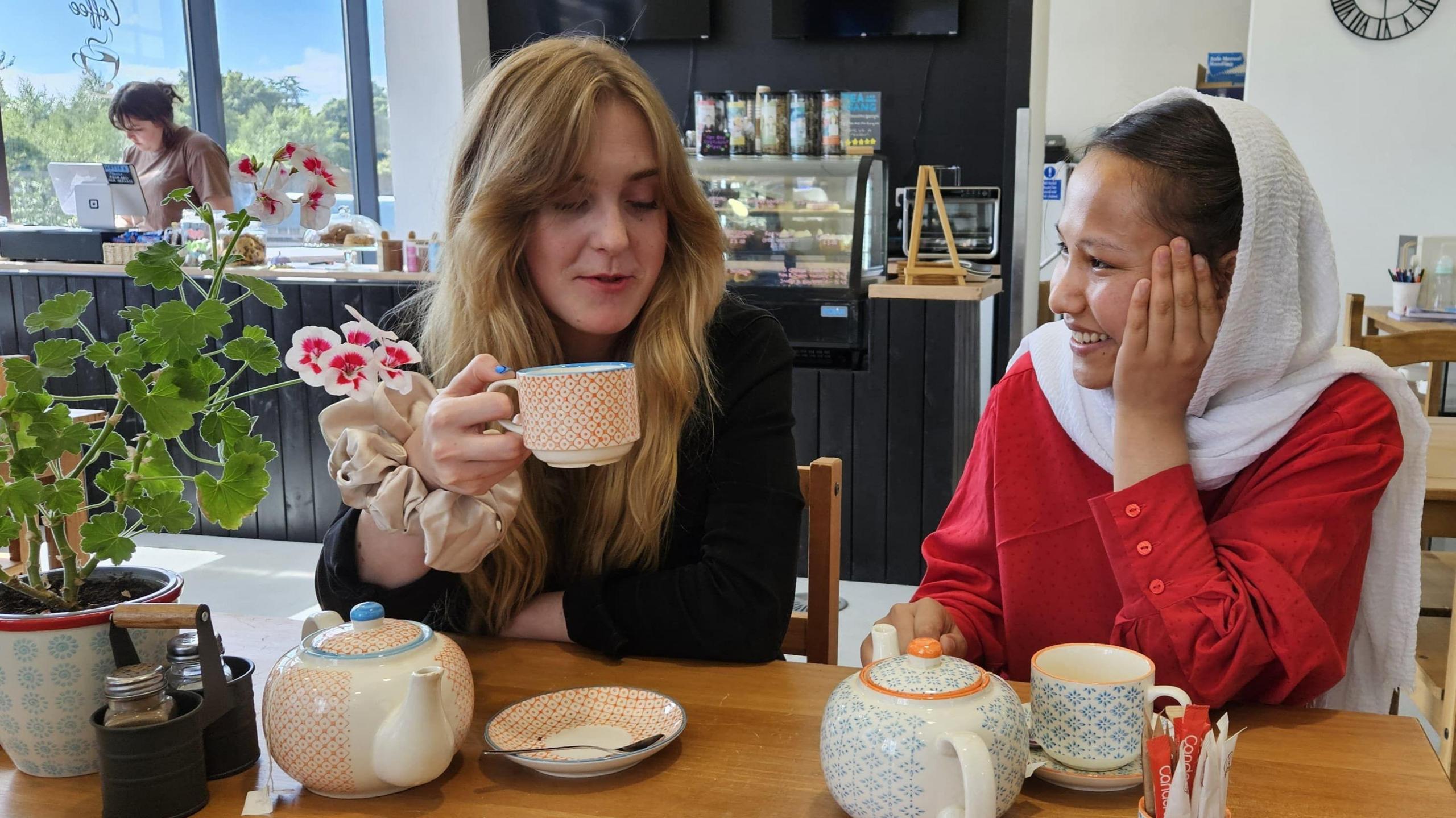 Eva Kearney and Zarifa Asghari sit together in a coffee shop. Eva is holding a mug, while Zarfia is looking at Eva