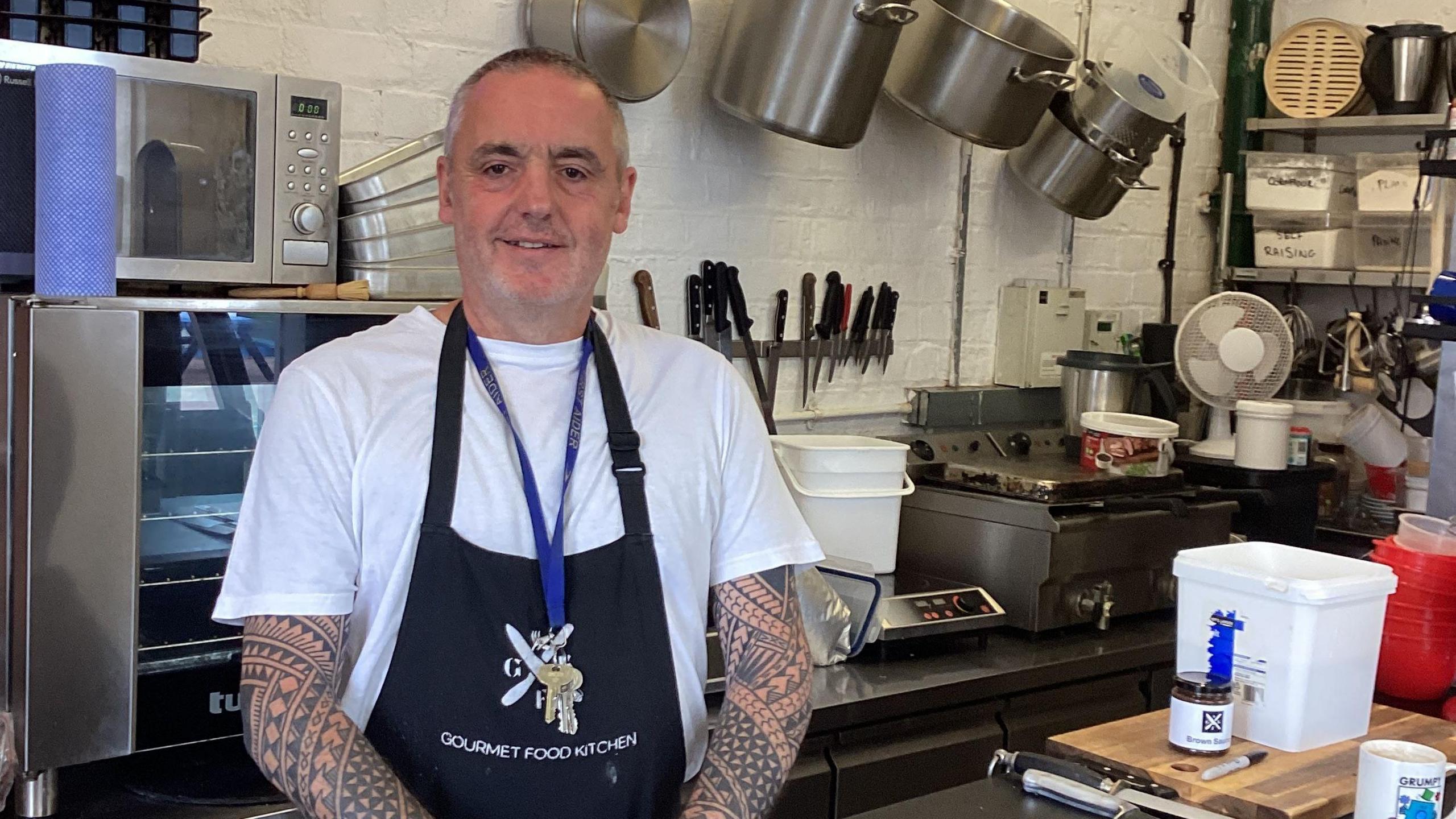 A man wearing a white t-shirt and a black apron stands in kitchen. Kitchen surfaces and equipment can be seen around him.