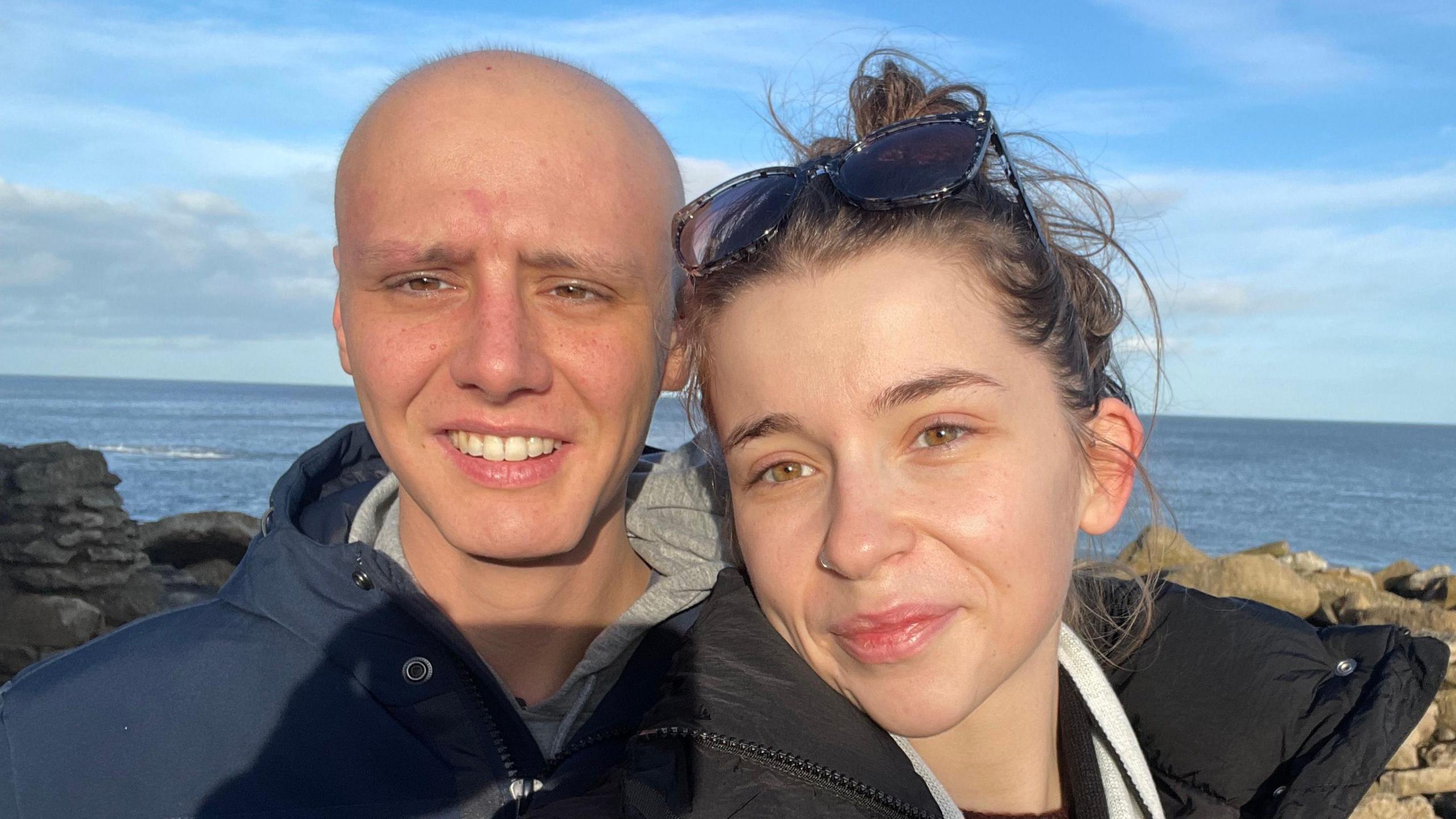 Josh, after his stem cell transplant, with his wife, Chloe. They're stood in front of a sea view. Josh has no hair.