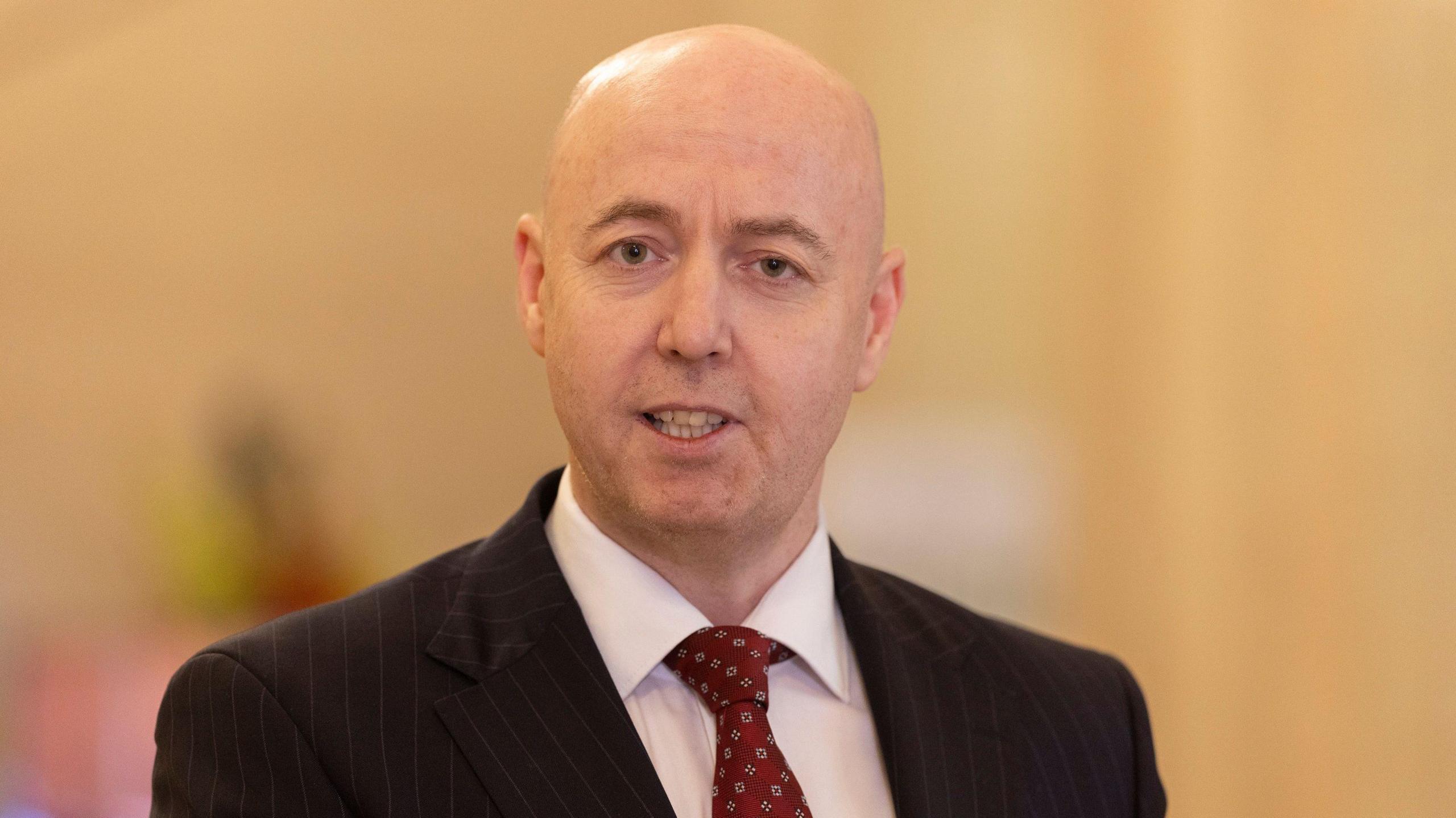 Jon Burrows is photographed looking toward the camera while speaking. He is bald and wearing a dark pinstriped suit, white shirt and red tie.