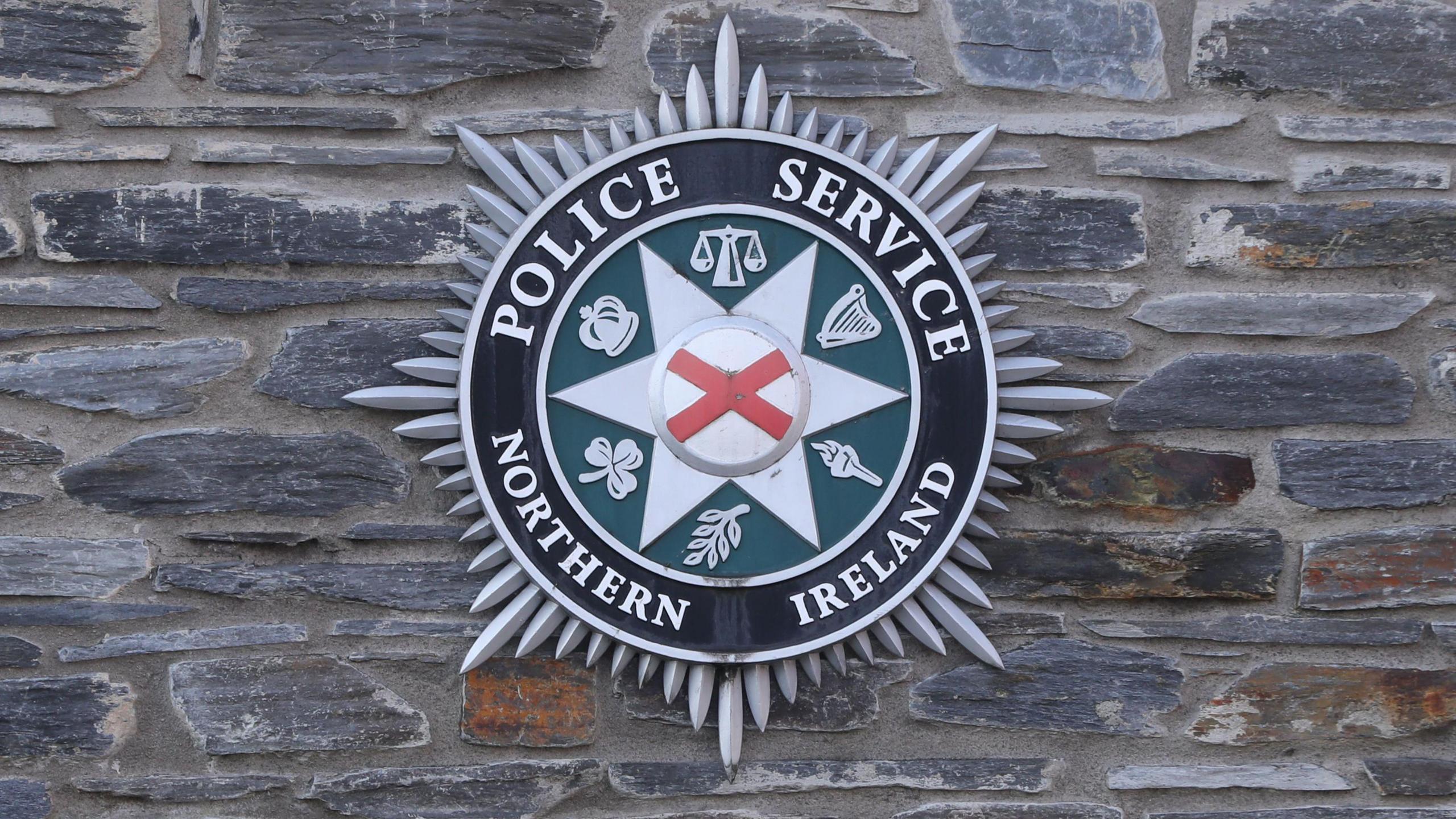 The PSNI badge on a stone wall. The badge is a Bath star with lettering around the inside reading "police service of Northern Ireland". In the centre is a red and white St Patrick's Saltire.